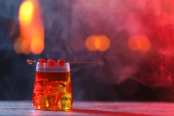 Ein Glas mit einem Rot-Getränk, Dekoriert mit einer Cocktailkirsche auf einem Cocktailspieß, vor einem unscharfen, bunten Hintergrund.