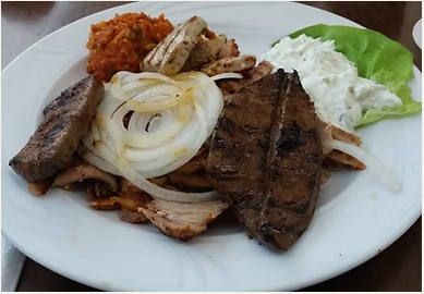 Ein Teller mit Grillfleisch, Zwiebeln, einer Portion Süßkartoffelpüree, einem Stück gegrillter Banane, Salatblättern und Weißdressing.