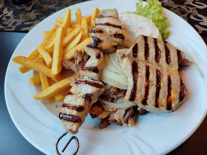 Auf einem weißen Teller sind gegrillte Fleischspieße, Pommes Frites, Salat mit Gurken, Tomaten und Zwiebeln, sowie ein Löffel Joghurt-Dip.