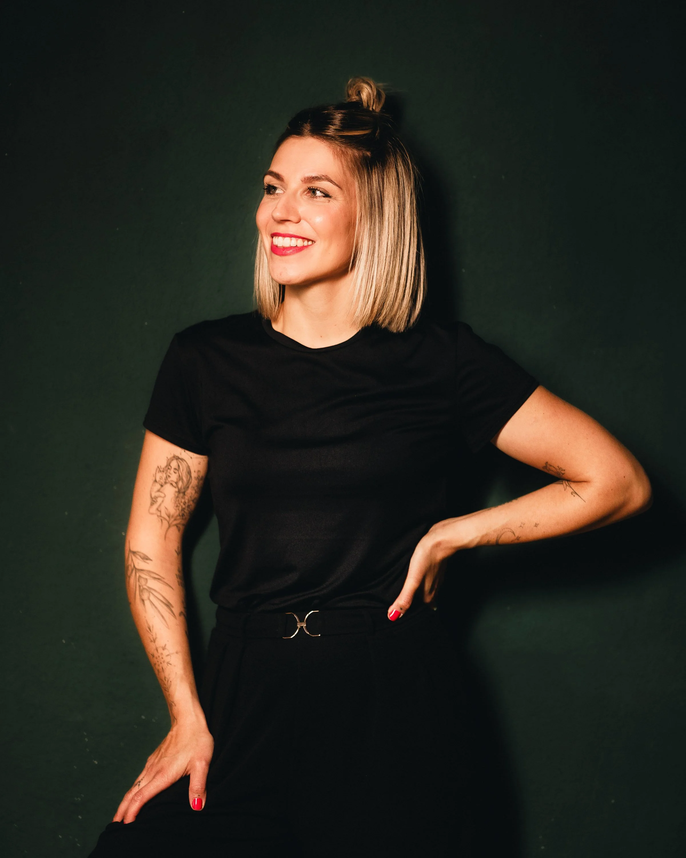 Eine lächelnde junge Frau mit Tattoo-Armen trägt ein schwarzes T-Shirt und schwarze Hosen vor einem dunklen Hintergrund.