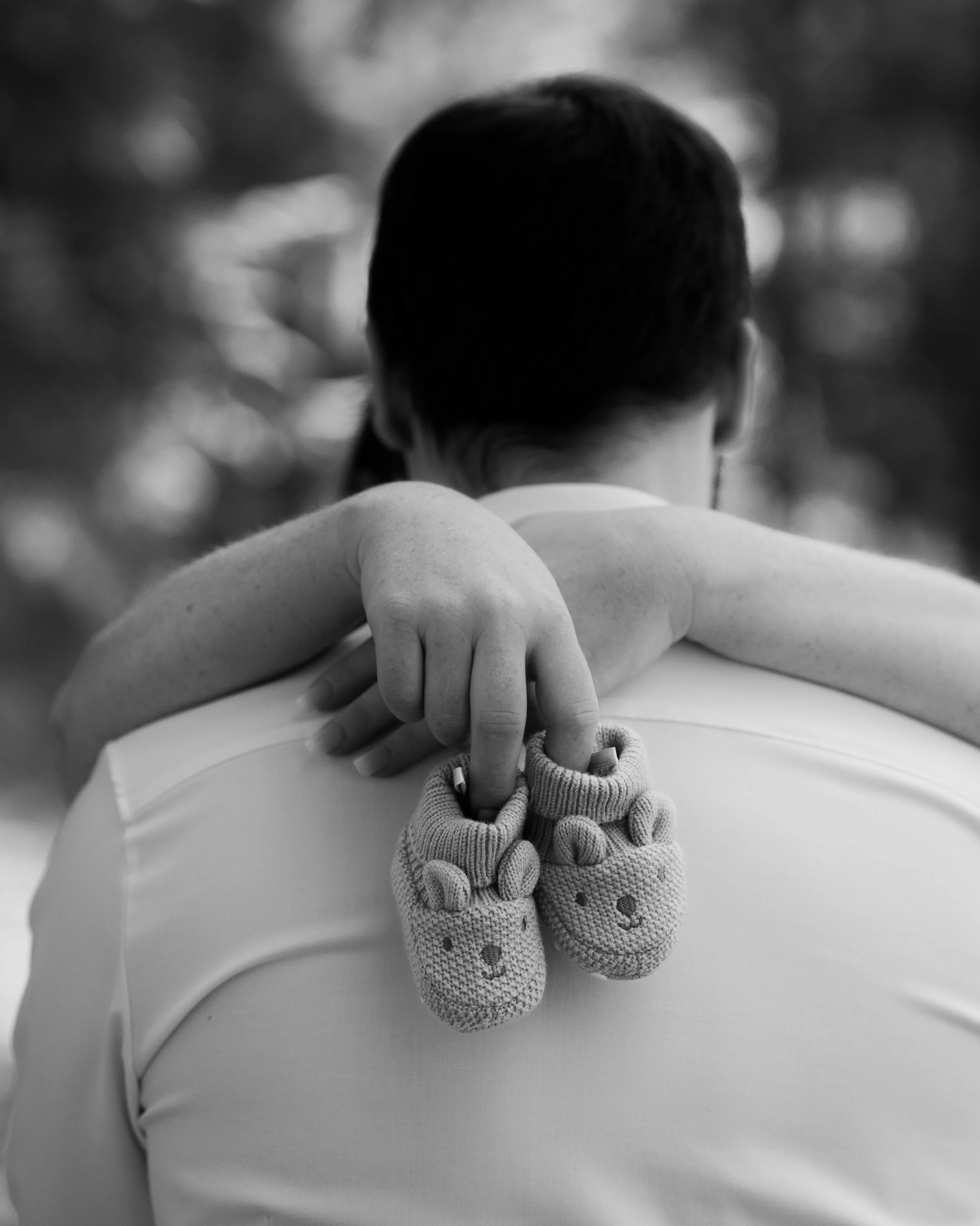 Ein Mensch mit kurzen dunklen Haaren sieht auf eine Frau mit gestrickten Babysocken. Die Person vor ihr trägt ein helles Oberteil, und die Szene ist in Schwarz-Weiß aufgenommen.
