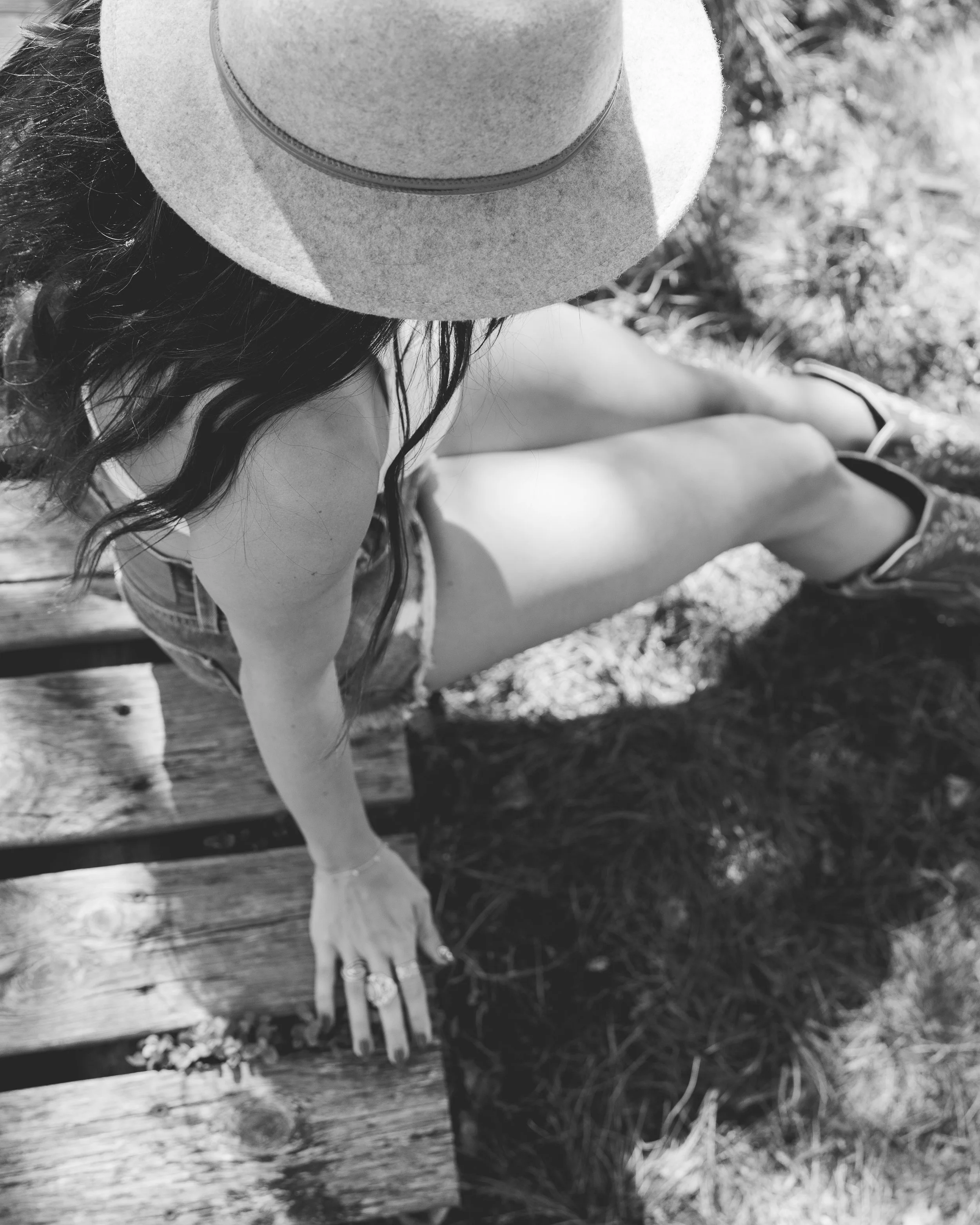 Junge Frau mit Sonnenhut, Sommerkleid und Flip-Flops sitzt auf einem Holzbalken im Freien, vermutlich im Park oder Garten, auf einem Sonnenbad, das Bild ist in Schwarz-Weiß gehalten.