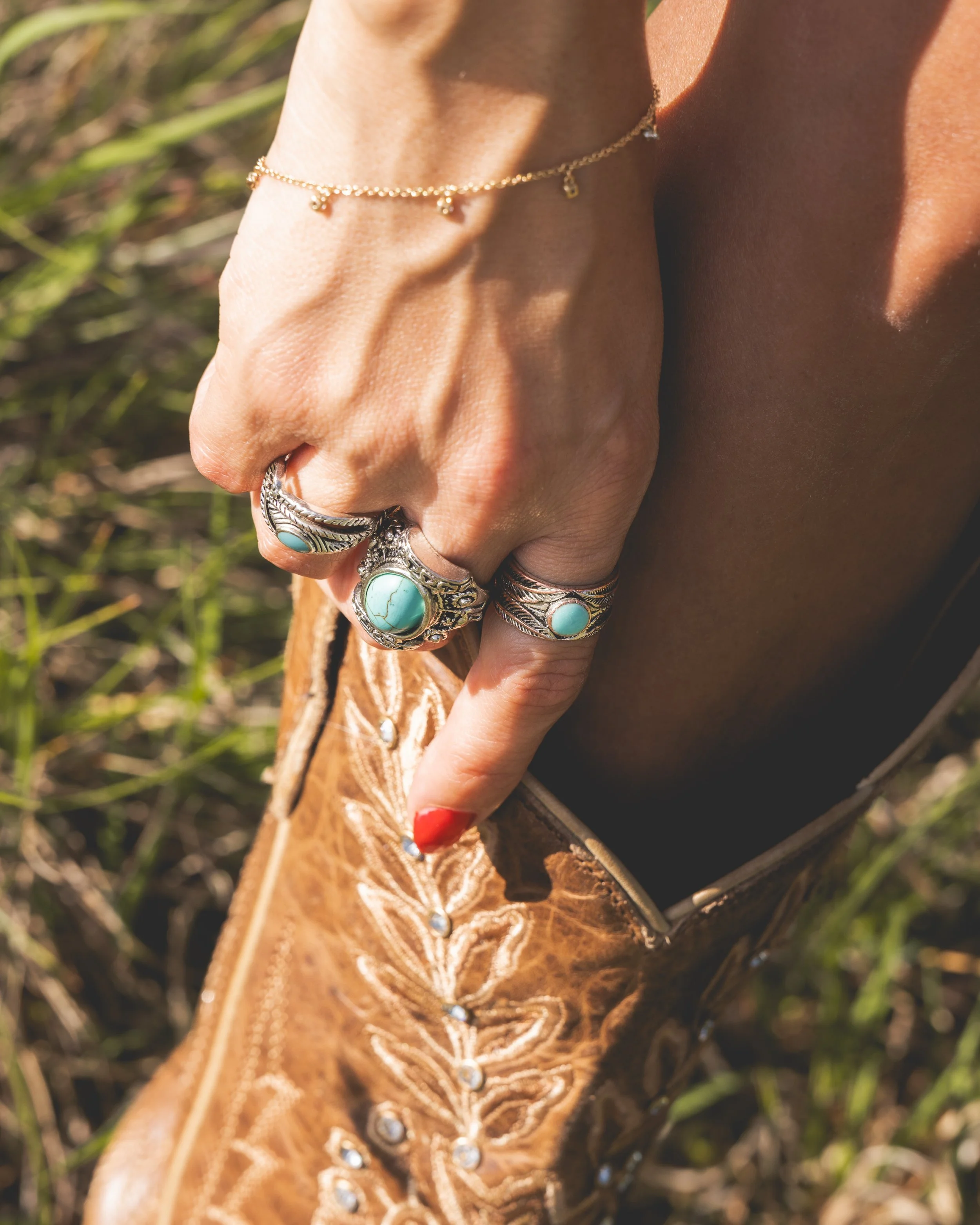 Nahaufnahme einer Hand mit mehreren silbernen Ringen, einer goldenen Armband mit kleinen Anhängern und bunter Nagellack, im Hintergrund grünes Gras.