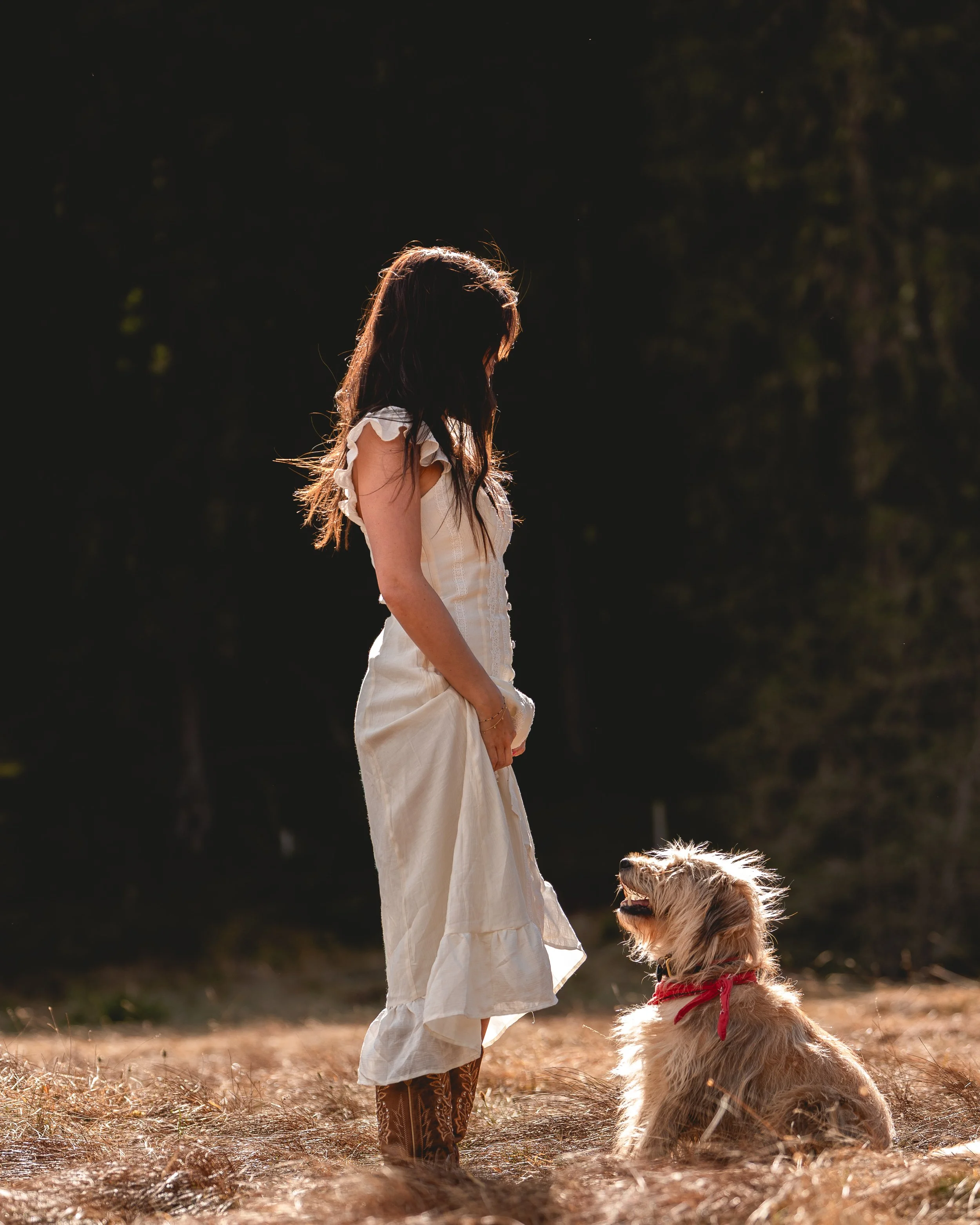 Ein Mädchen mit langen Haaren trägt ein weißes Kleid und steht auf einer Wiese, während ein Hund mit Rotem Halstuch sitzt und auf sie blickt.