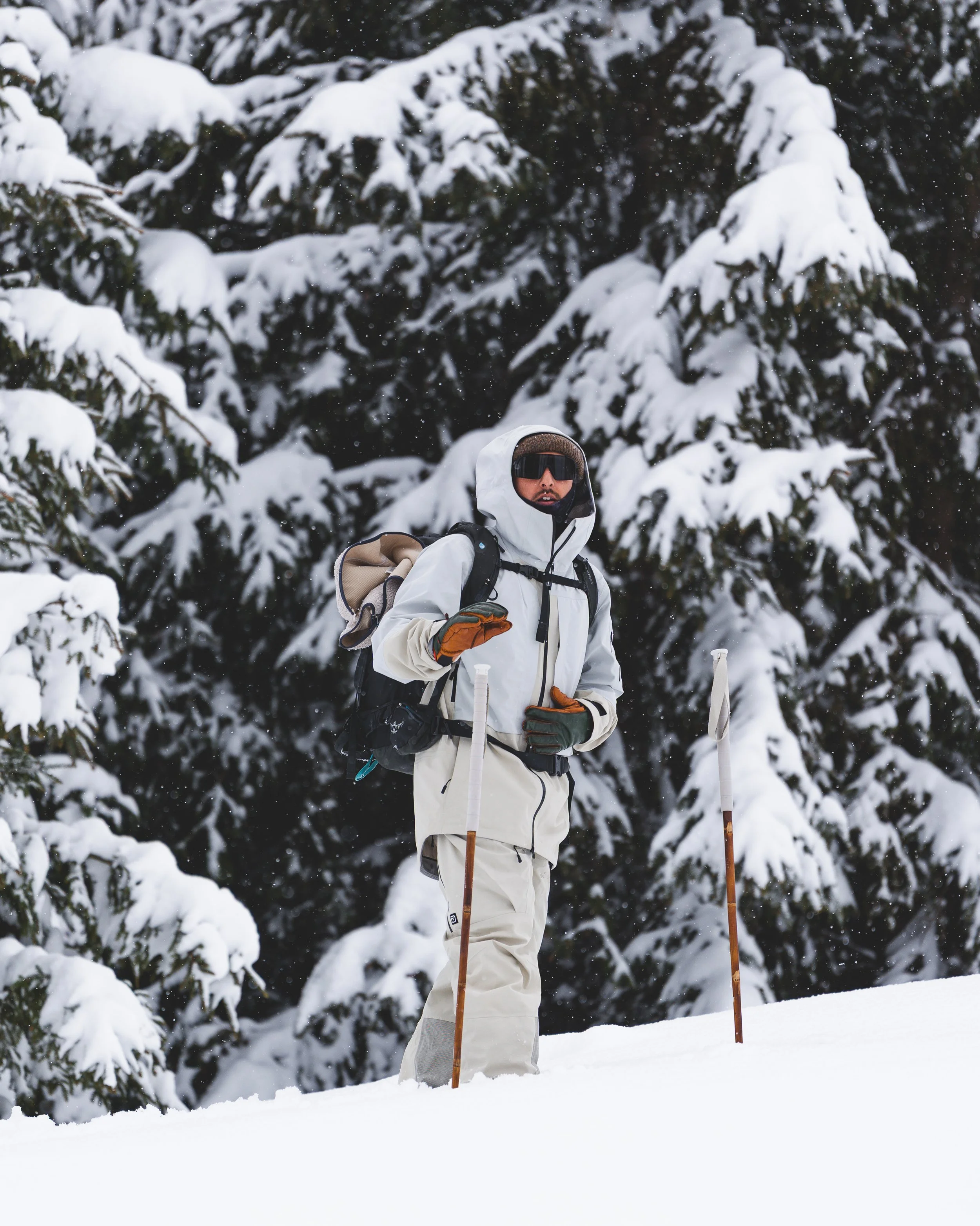 Ein Mensch trägt Winterbekleidung, eine weiße Jacke und eine Kapuze, in einer verschneiten Waldlandschaft und hält Wanderstöcke in den Händen.