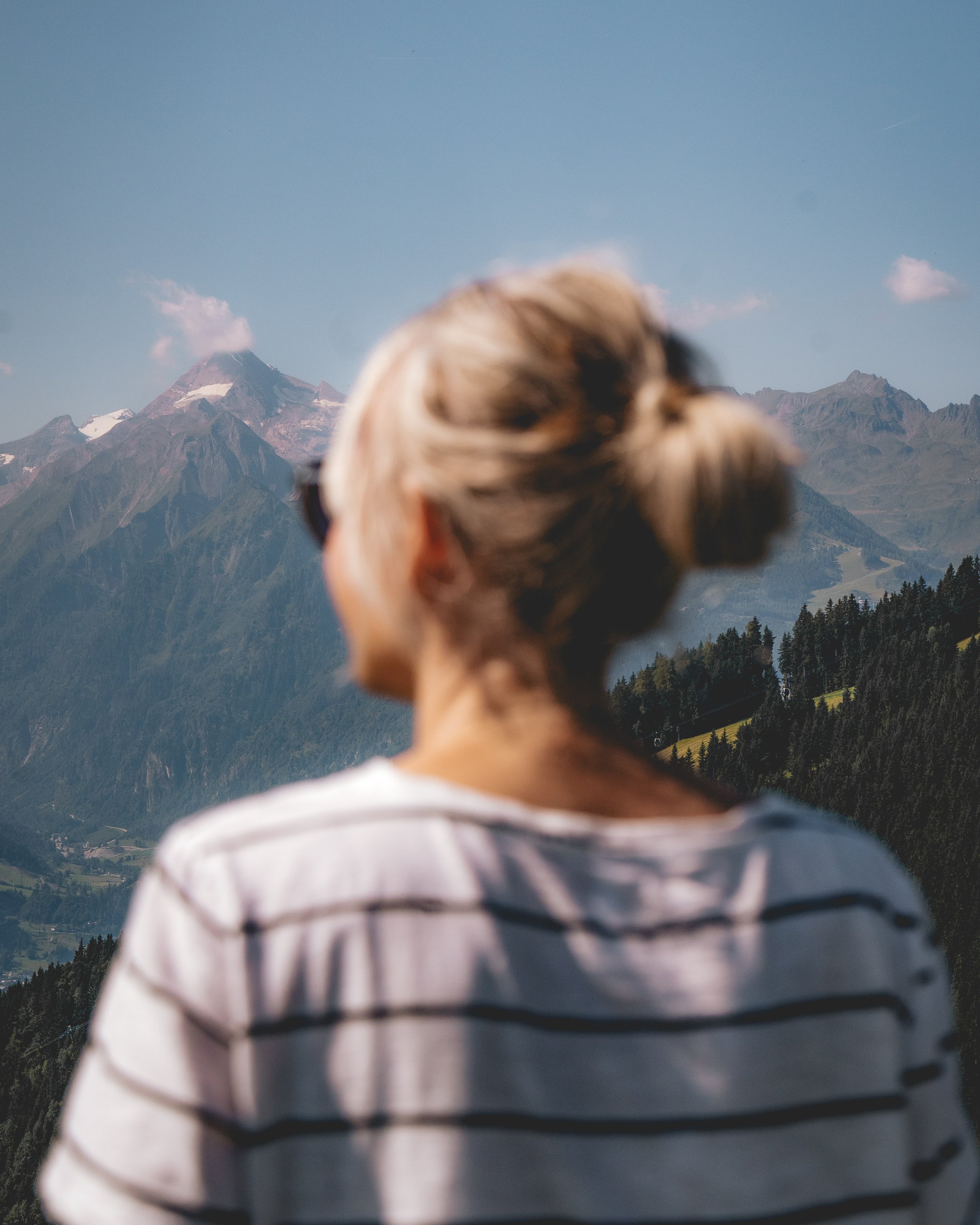 Eine Person mit einem Dutt trägt ein gestreiftes Hemd und blickt auf die Berge im Hintergrund, darunter einen Vulkan mit Schneeflecken und umliegende bewaldete Hügel, bei schönem Wetter.