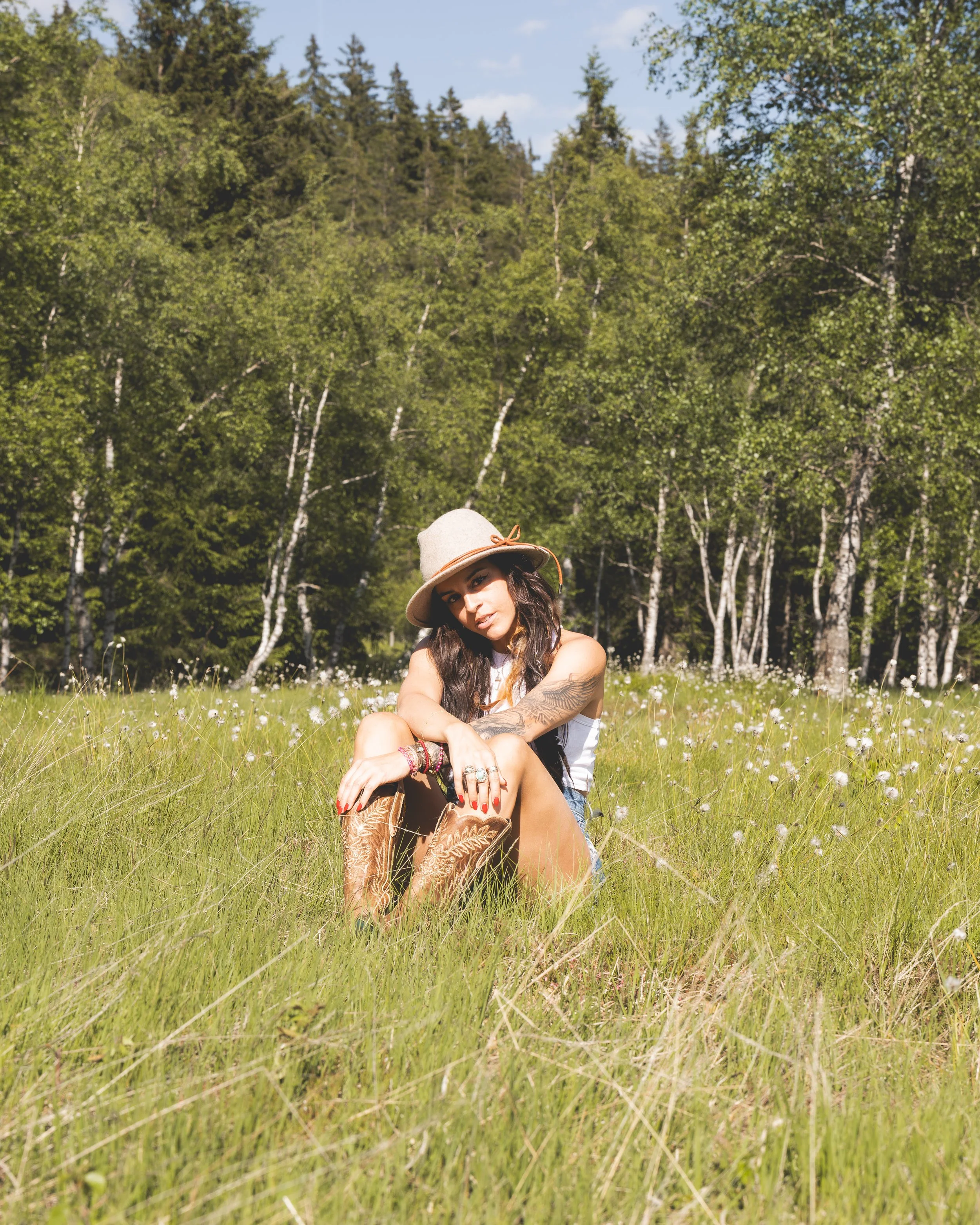 Frau sitzt im Gras in einem Feld mit weißen Blumen, trägt einen Hut und Blick in die Kamera, mit Bäumen im Hintergrund und blauem Himmel.