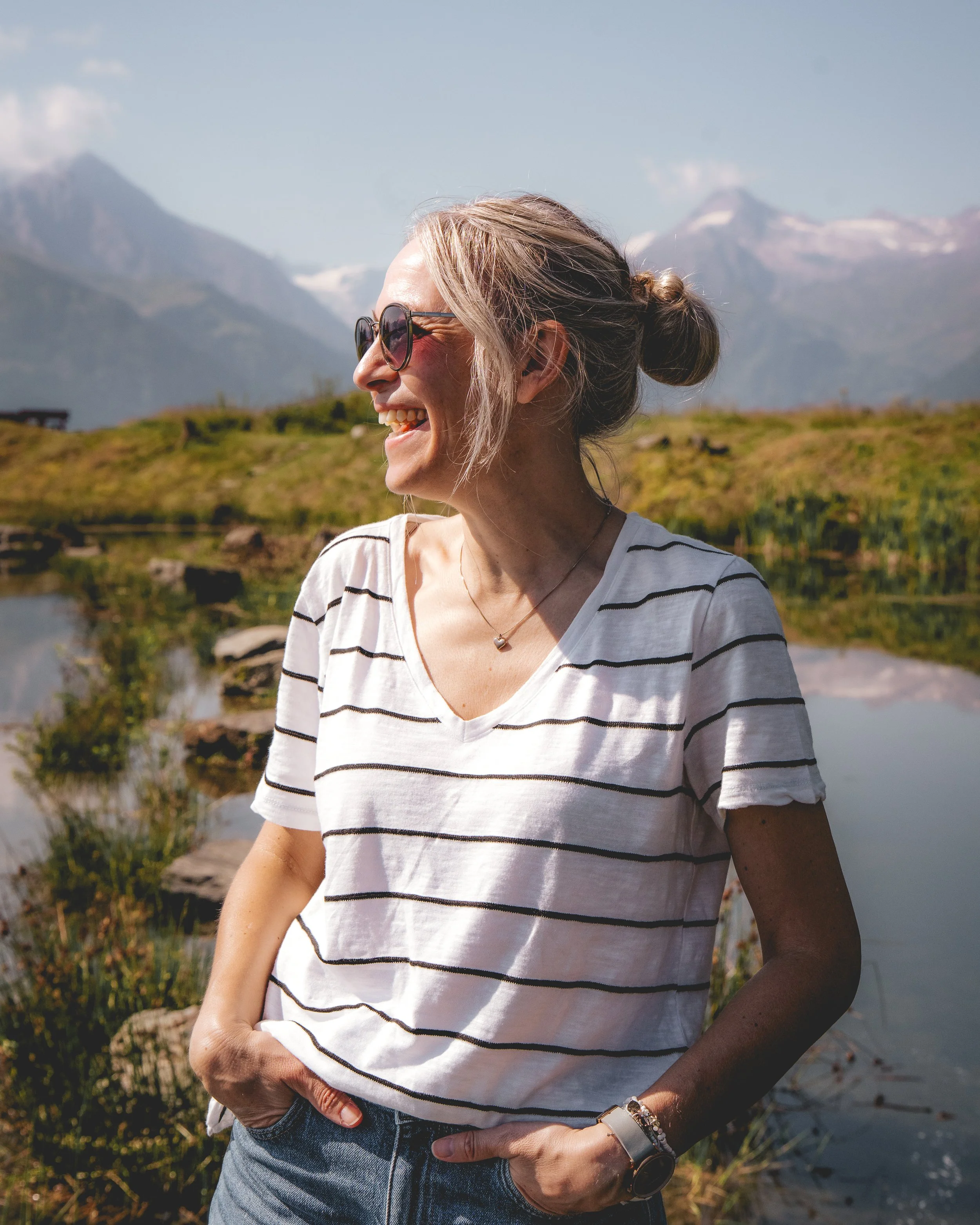 Eine lachende junge Frau mit Sonnenbrille, gestreiftem T-Shirt und Jeans vor einer bergigen Landschaft mit Gewässer im Hintergrund.