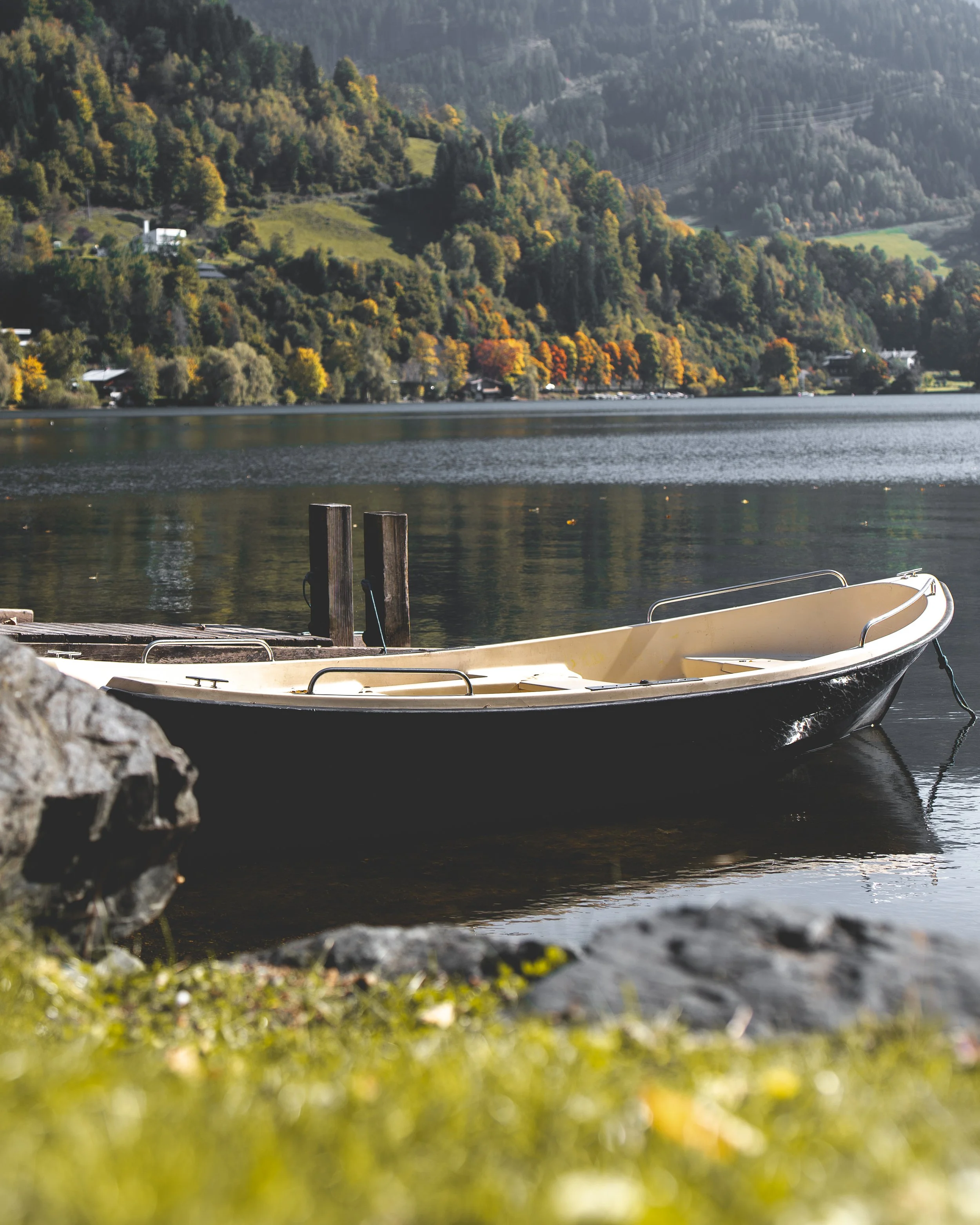 Ein Boot an einem Steg am See, mit Blick auf bewaldete Hügel und Berge im Hintergrund, im Herbst mit buntem Laub.