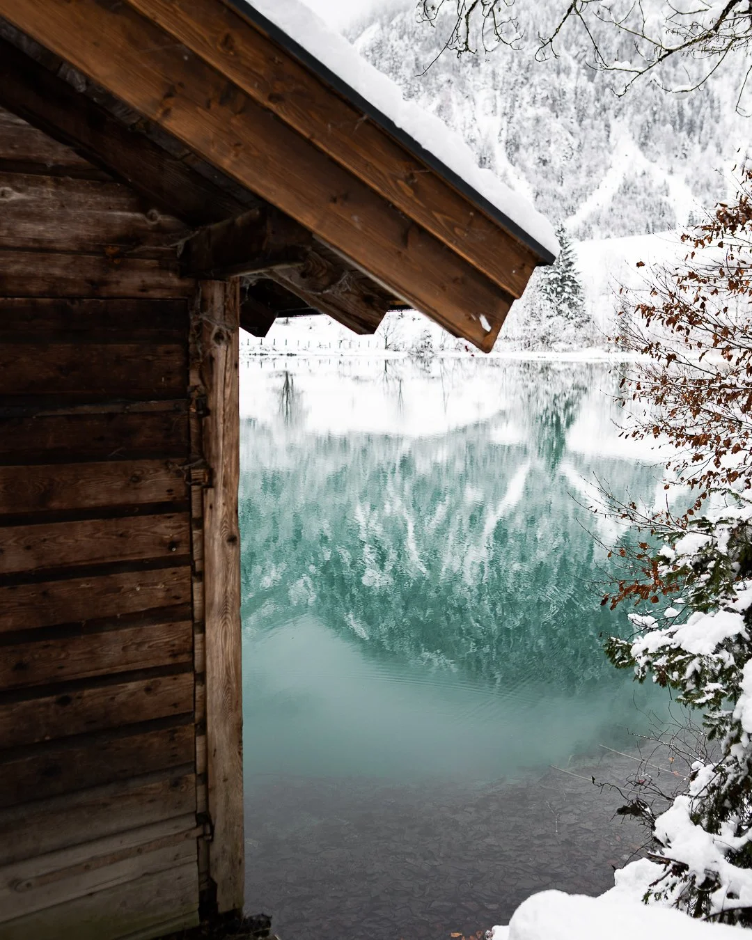 Holzhaus an einem verschneiten Ufer eines Sees mit schneebedecktem Wald im Hintergrund.