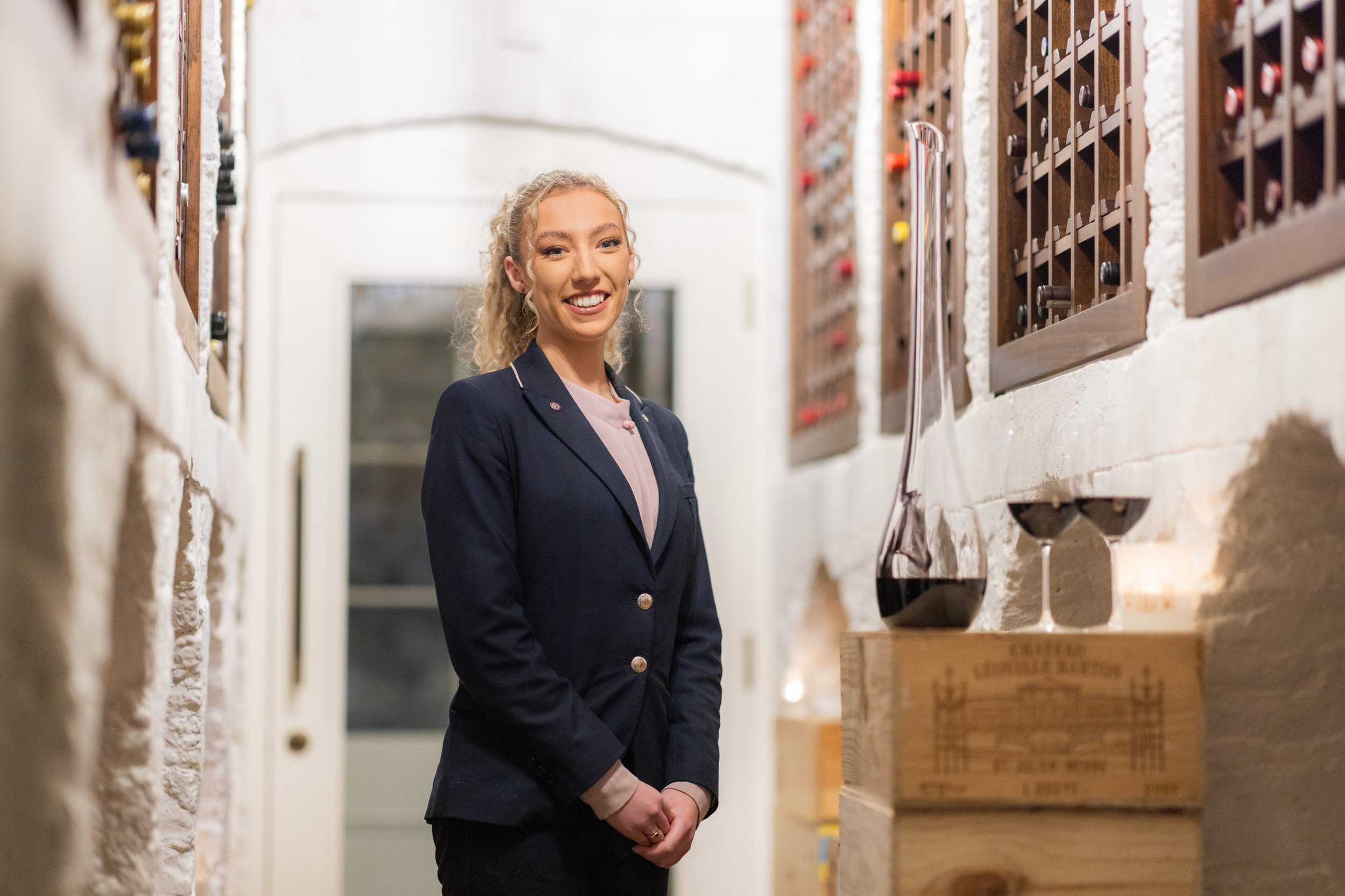 A smiling woman in a navy blazer and light pink blouse standing beside wine bottles and glasses in a wine cellar.