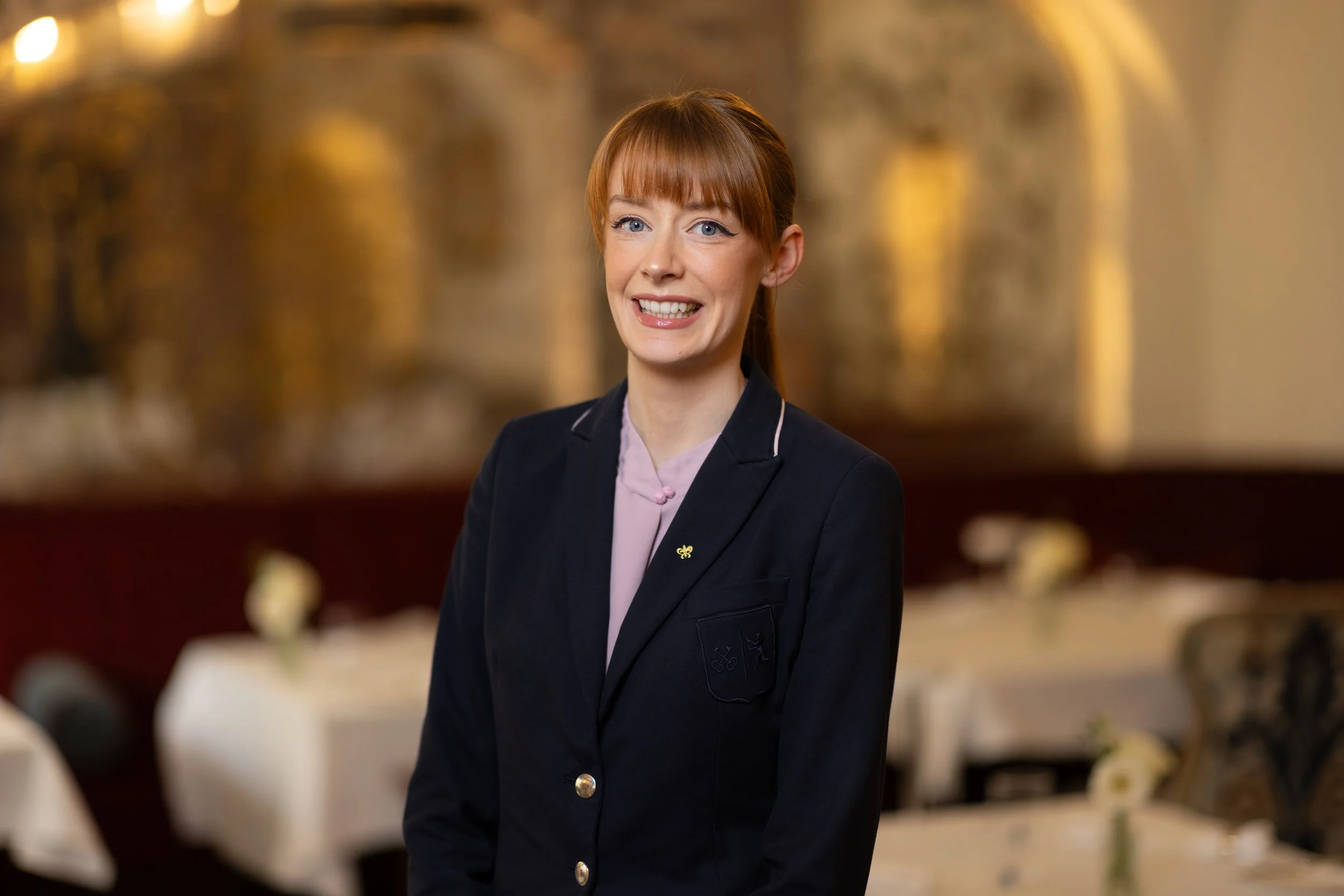 A woman with red hair and blue eyes smiling, dressed in a dark blazer with a light pink blouse, standing in an elegant setting with blurred background of a gold-colored sculpture or artwork.
