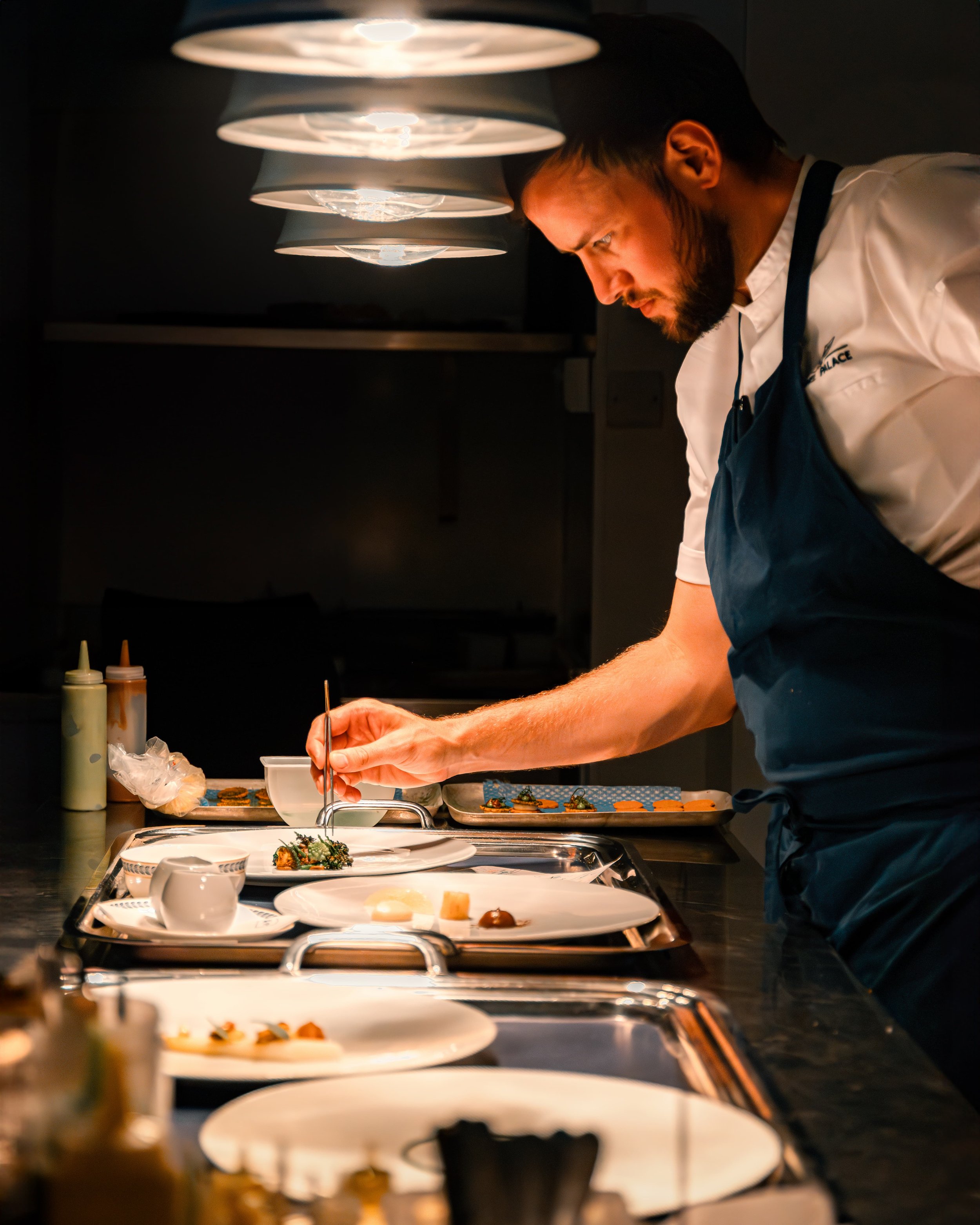A chef in a white uniform and blue apron is preparing food in a professional kitchen, illuminated by overhead lights, with various plates and condiments on the counter.