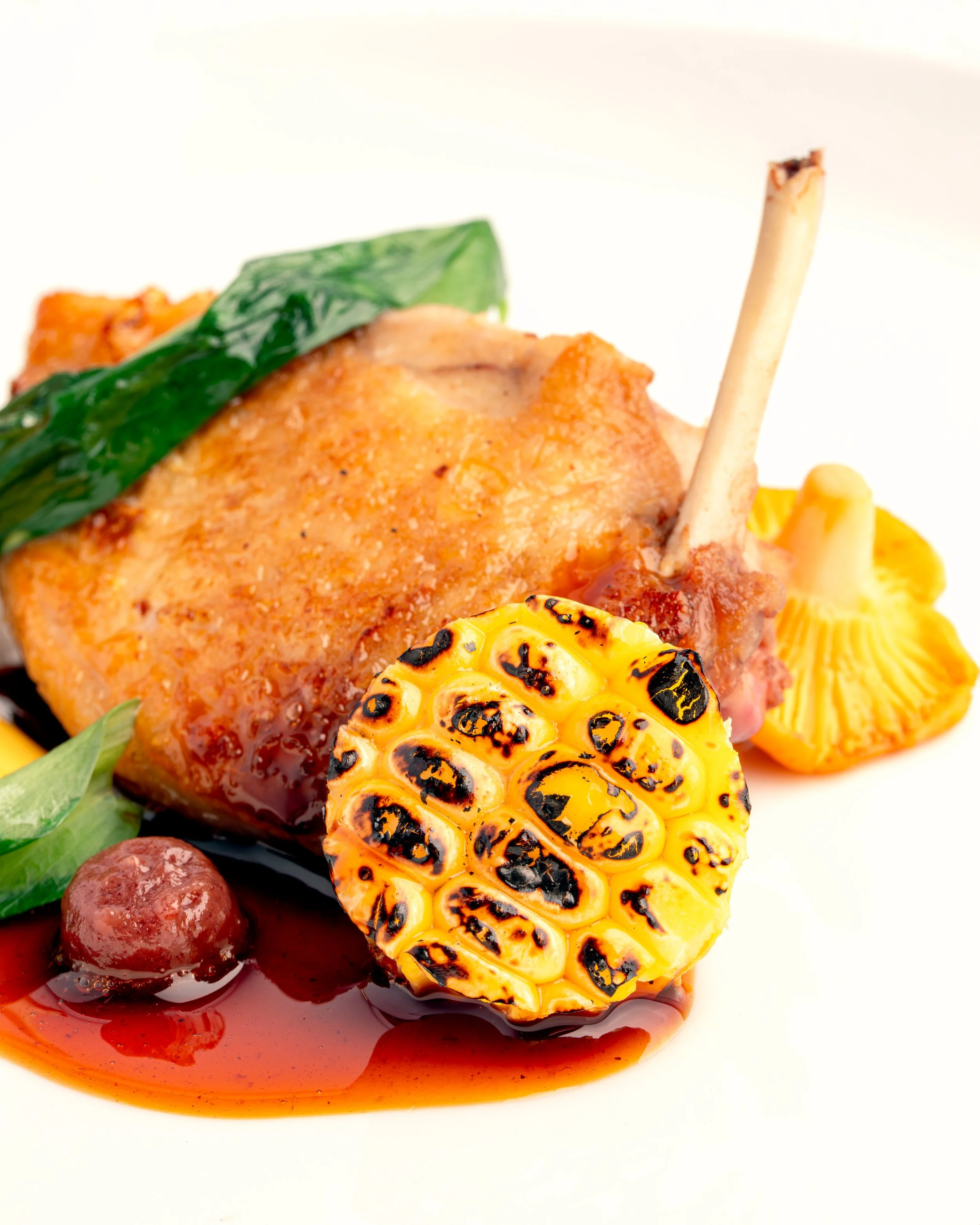 Close-up of grilled corn on the cob, roasted tomatoes, and a piece of grilled chicken with herbs, served with a dark sauce and garnished with grilled pepper and fresh green leaves.