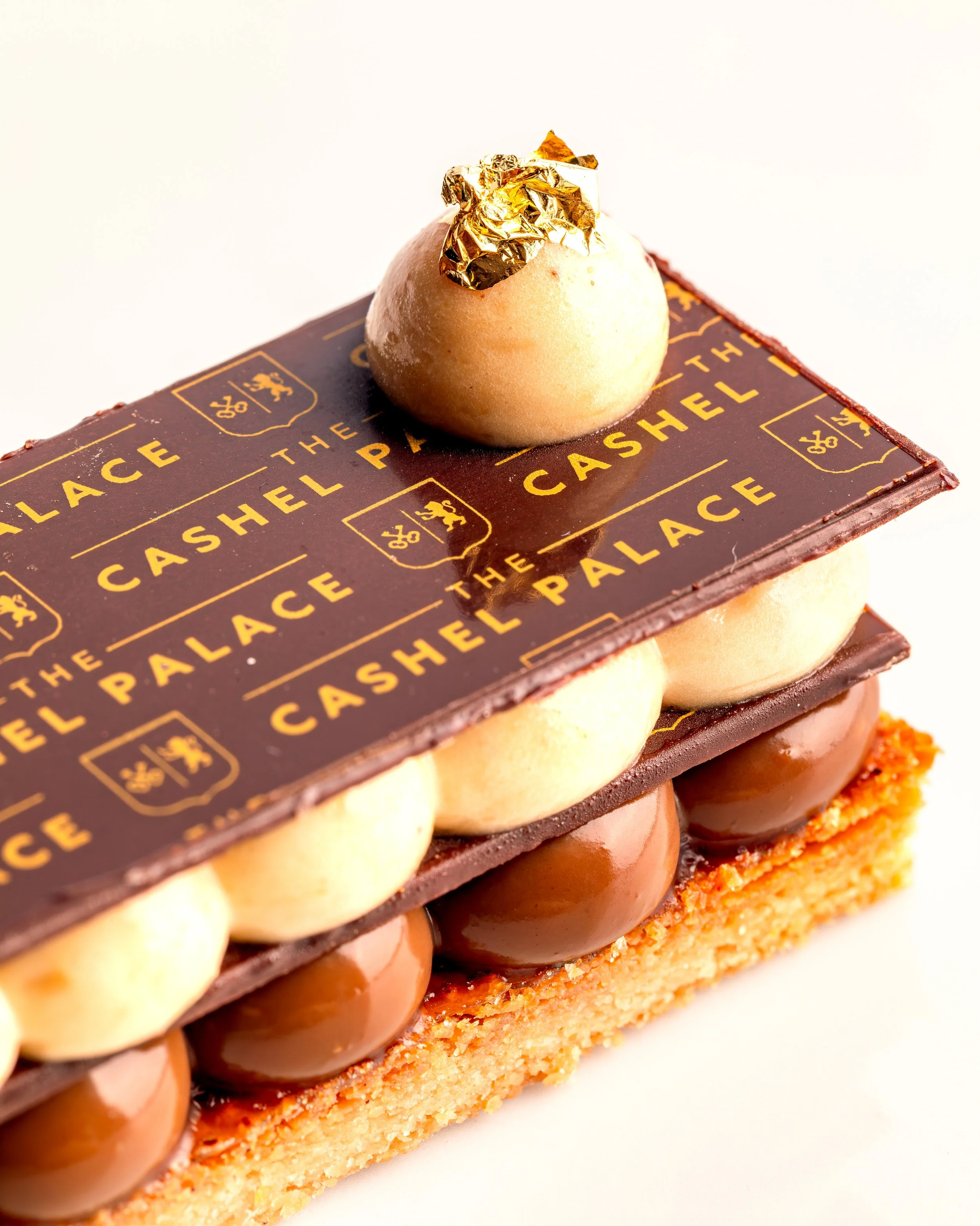 A layered pastry dessert with a base of crumbly golden crust, topped with a row of glossy chocolate balls, and topped with a small dome of mousse garnished with a piece of gold leaf.