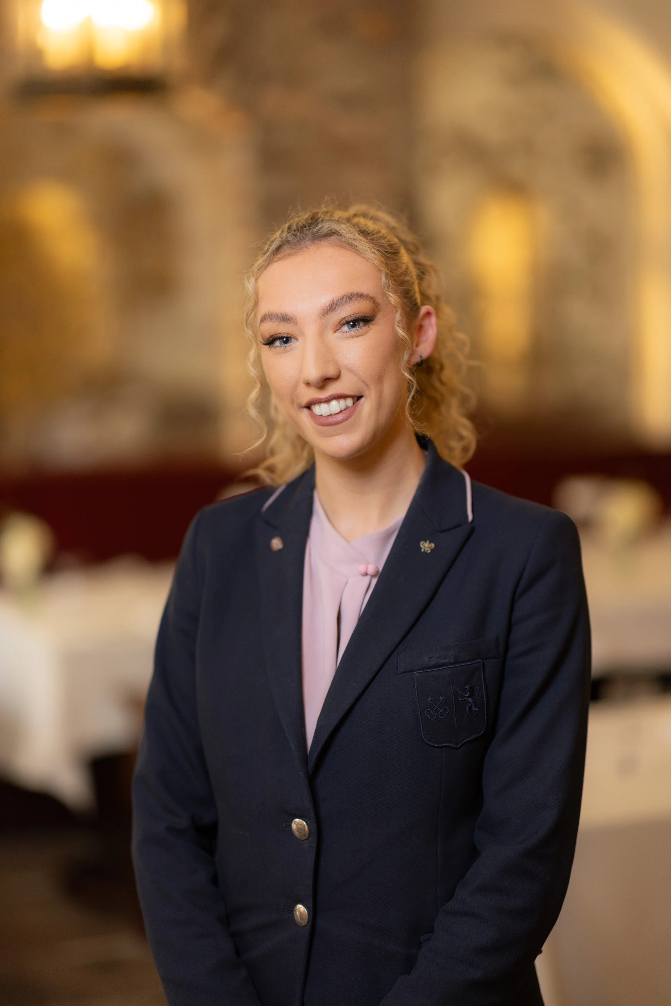A smiling young woman with curly blonde hair and blue eyes wearing a navy blazer and a light pink blouse, standing indoors with shelves of various objects blurred in the background.