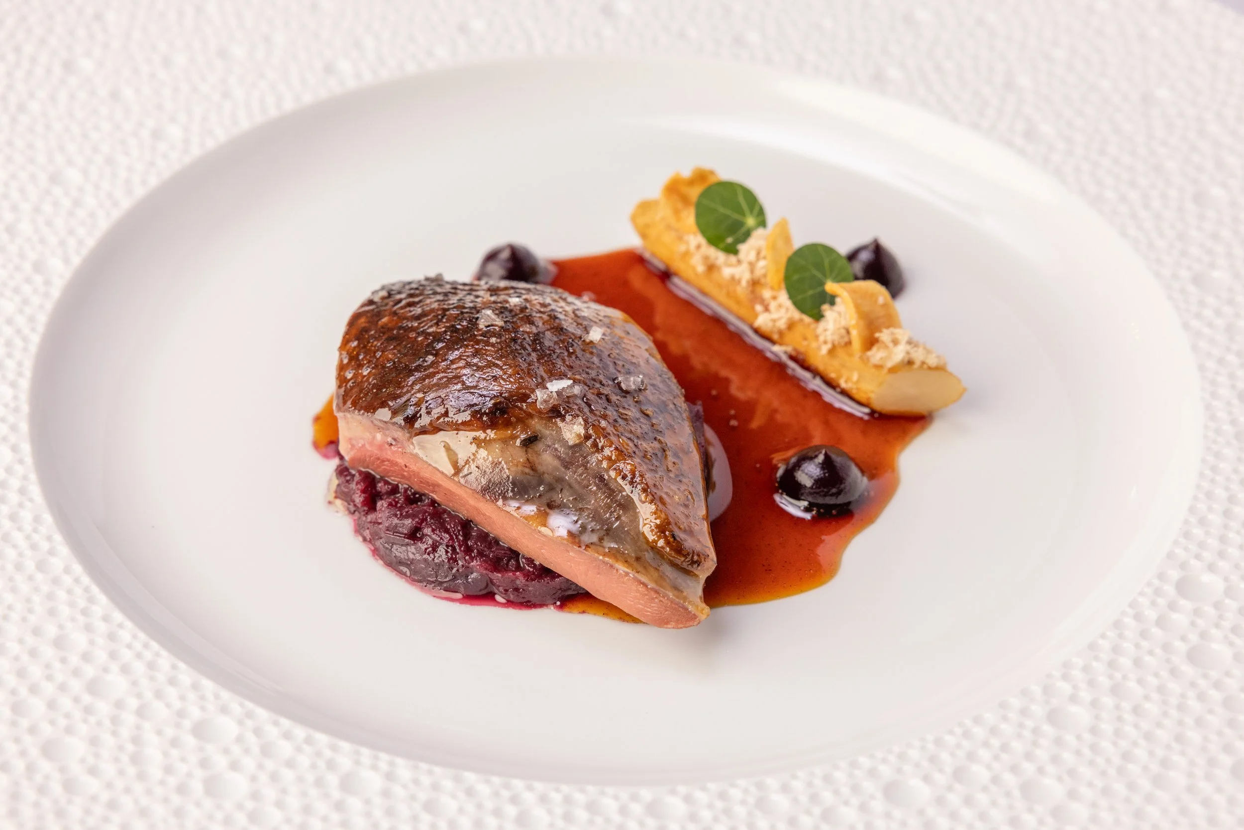 A plated gourmet dish featuring a piece of cooked meat with a crispy skin, served with a red sauce and garnished with edible greens and yellow decorations.
