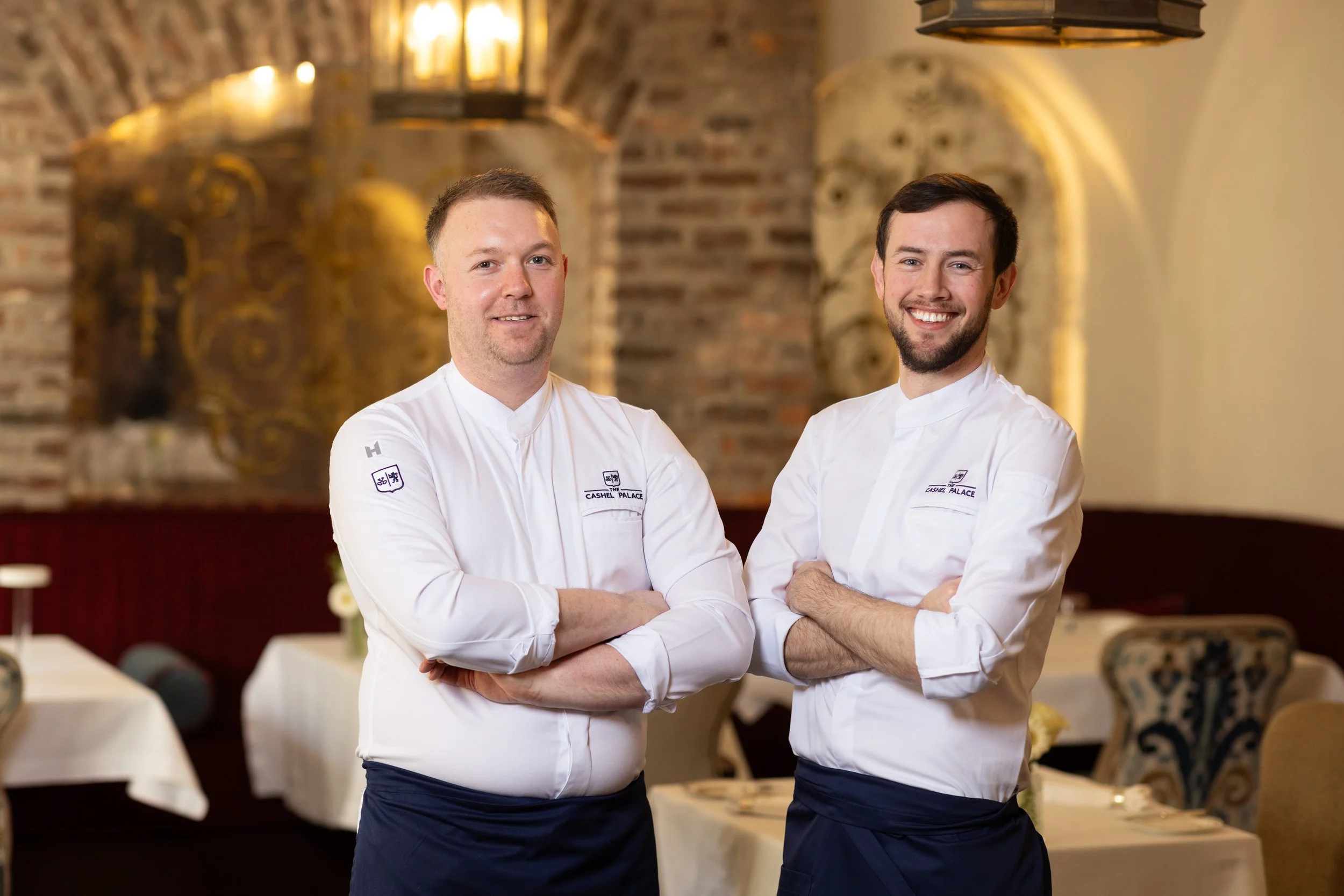 Two male chefs in white uniforms with arms crossed, standing in a restaurant with brick walls and hanging lights.
