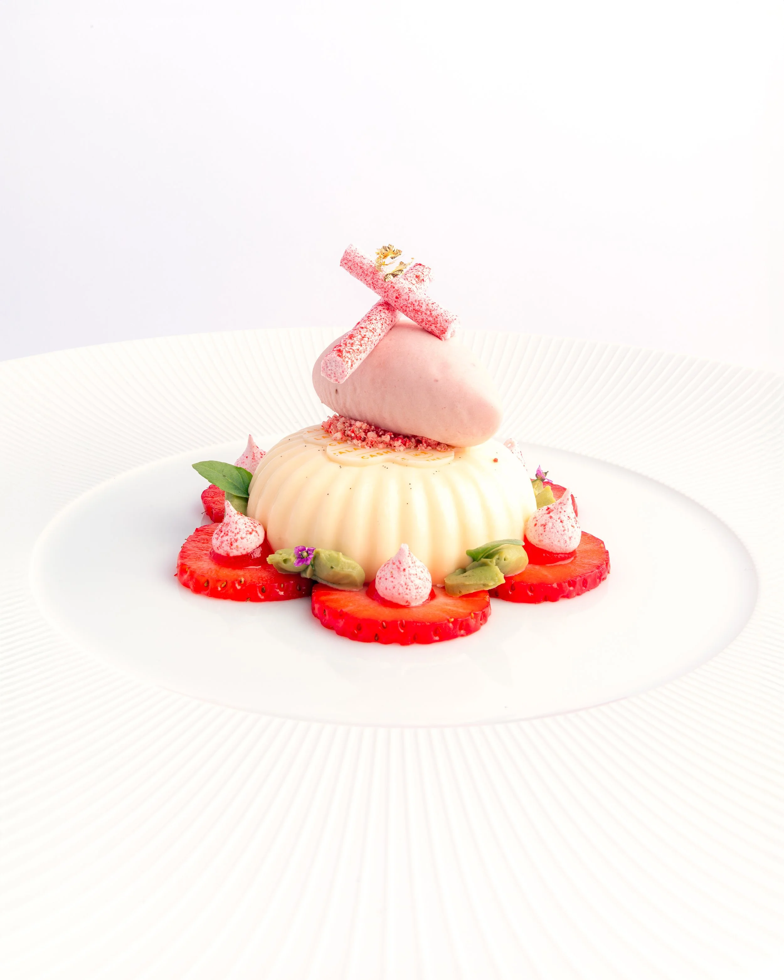 A dessert with a pink mousse heart topped with a pink rolled candy and small edible flowers on a white plate with a decorative border.