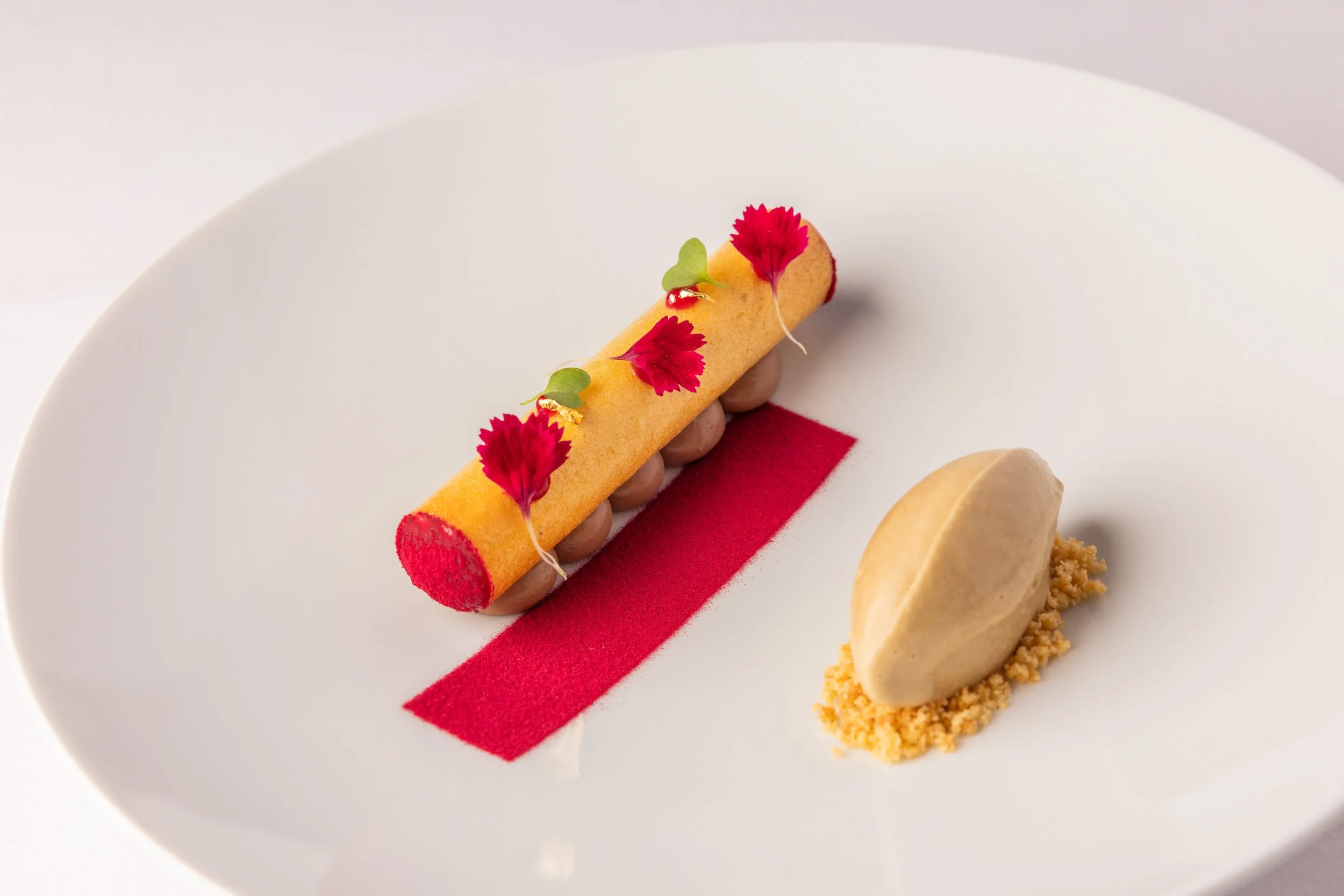Elegant plated dessert featuring a cylindrical yellow roll decorated with red edible flowers and small edible leaves, placed on a white plate with a red rectangular decoration and accompanied by a scoop of beige ice cream on a crumb-like base.