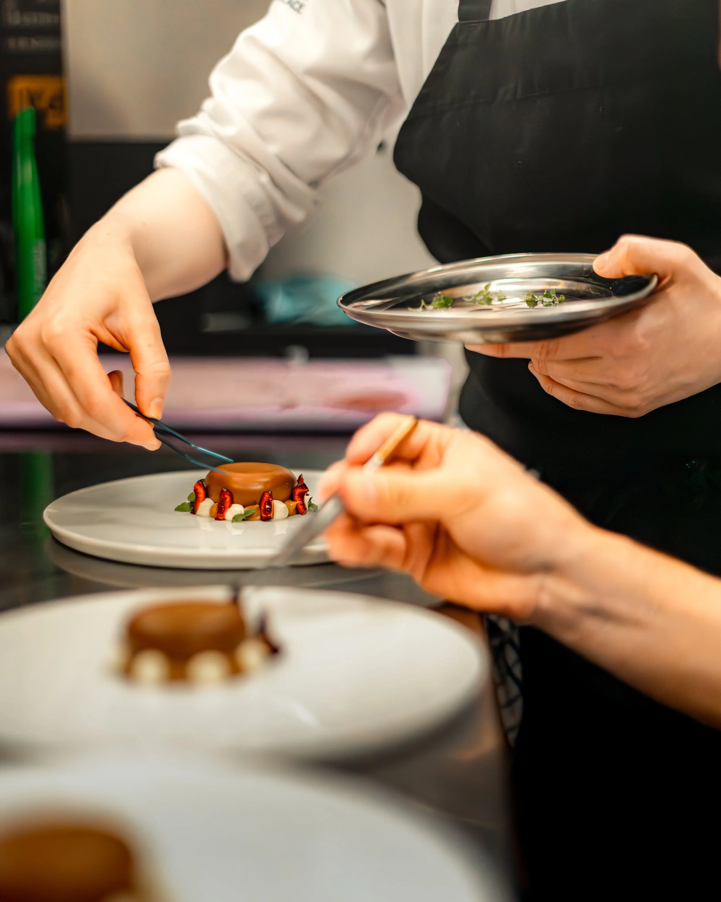 Close-up of a chef decorating a small, round cake with strawberries and white cream on a white plate, with another person holding a piping bag in the foreground.