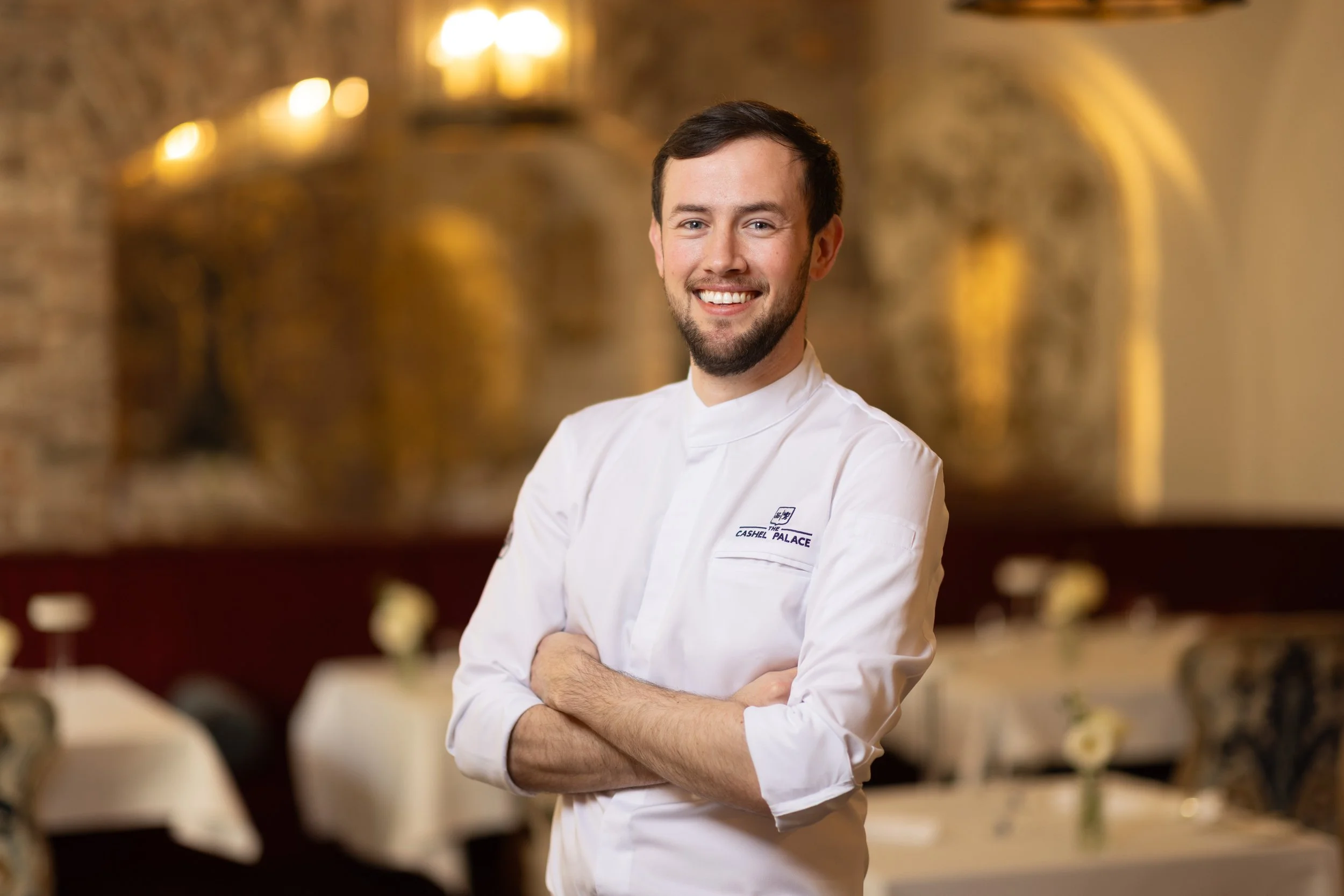 A smiling male chef with a beard and dark hair, wearing a white chef's coat, standing in a warmly lit restaurant or kitchen setting with blurred background of tables and chairs.