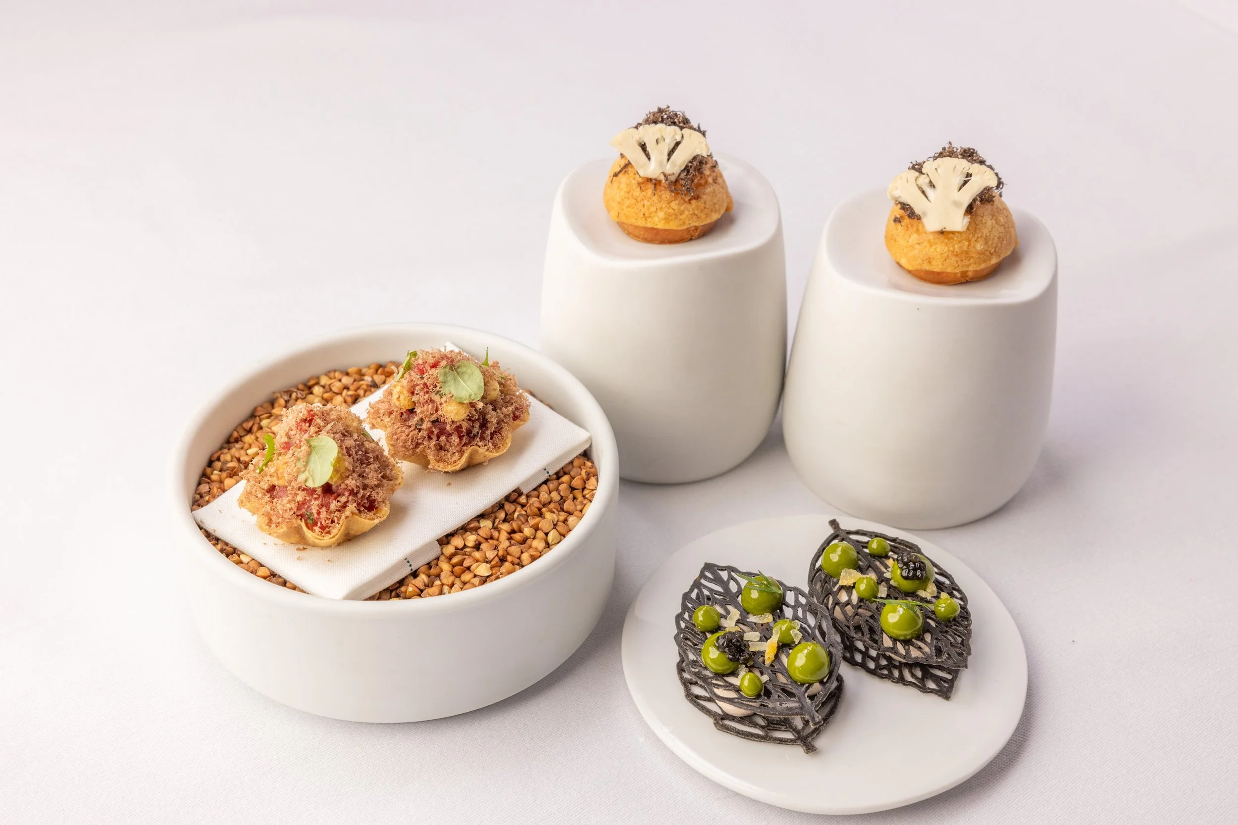 Assorted gourmet desserts on white dishes and stands, including small chocolate lattice topped with green and black berries, a bowl with topped fried pastries garnished with microgreens, and small pastries with white chocolate decorations.