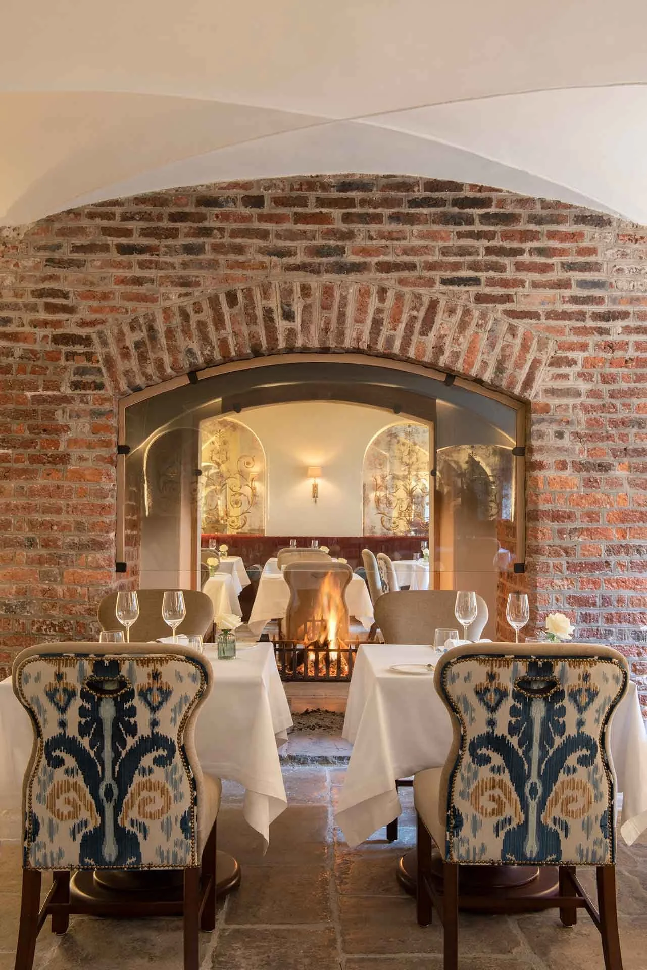 Elegant restaurant dining area with tables set with white tablecloths, wine glasses, and cutlery, featuring a brick archway with a fireplace and cozy lighting.