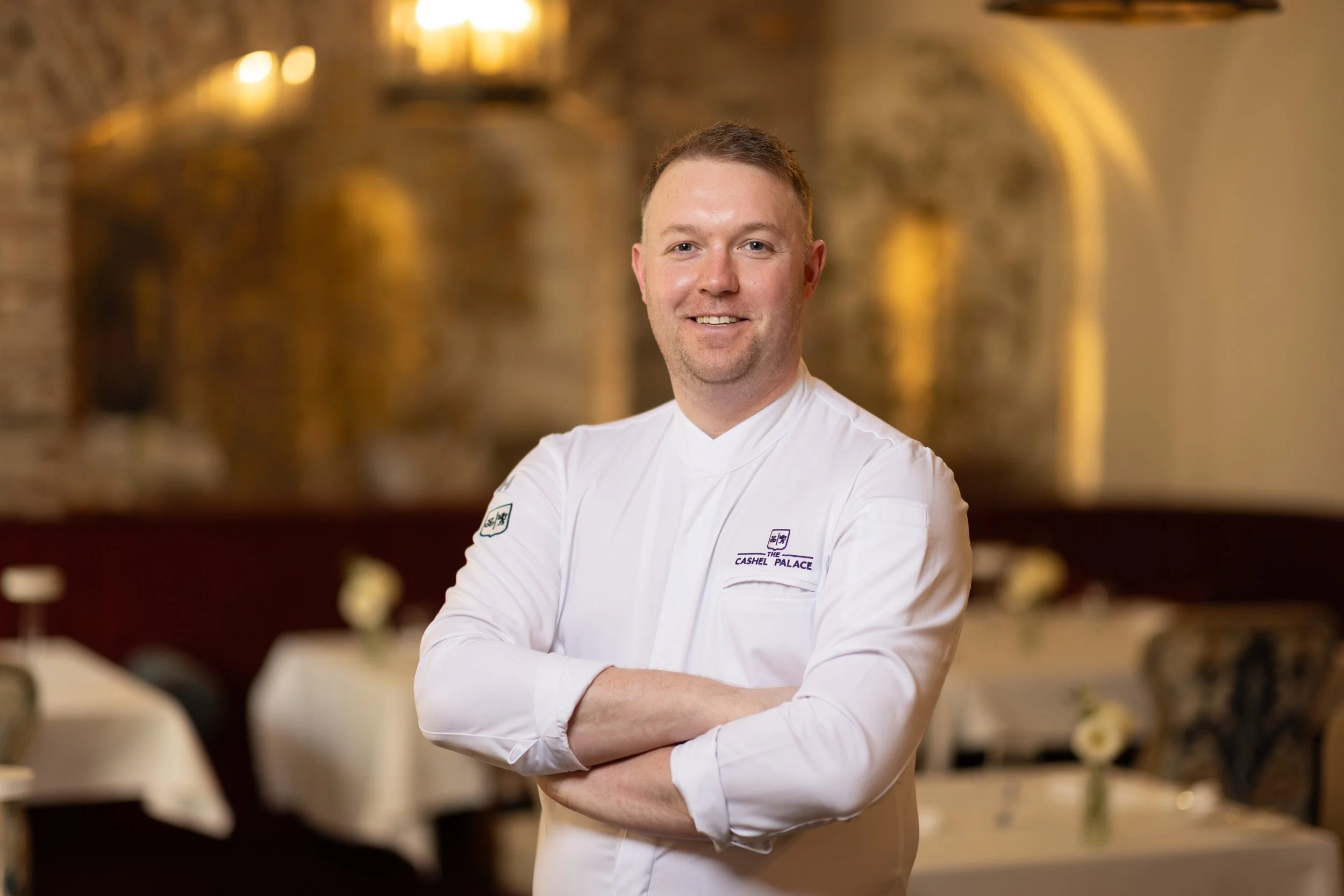 A male chef smiling with arms crossed in a restaurant setting with warm lighting and blurred tables and chairs in the background.