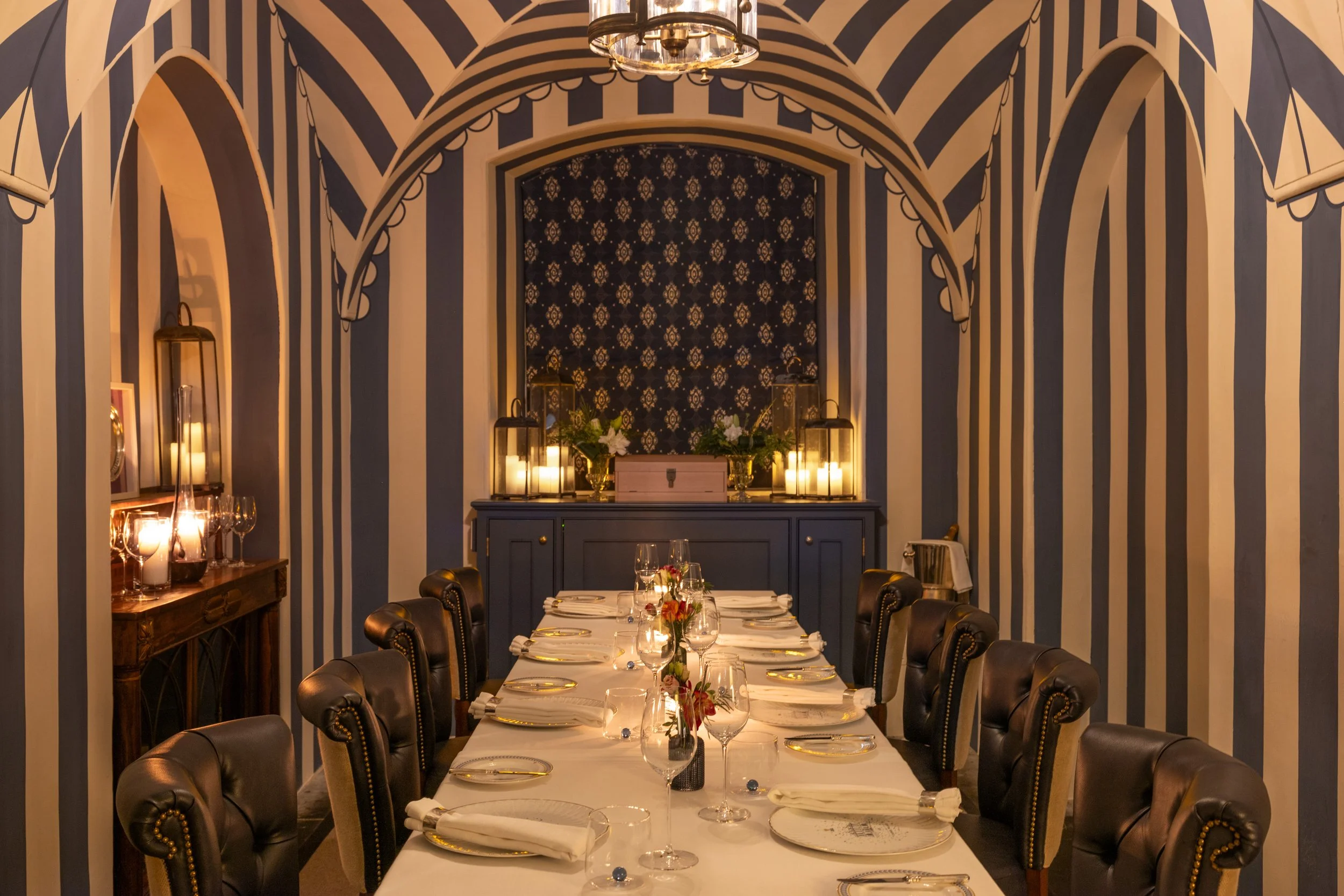 Elegant dining room with striped navy and white wallpaper, arched ceiling, and a long banquet table set for a formal dinner with white tablecloths, wine glasses, and floral centerpieces. Candles are lit along a sideboard, and there is a decorative wall with floral wallpaper and candles.