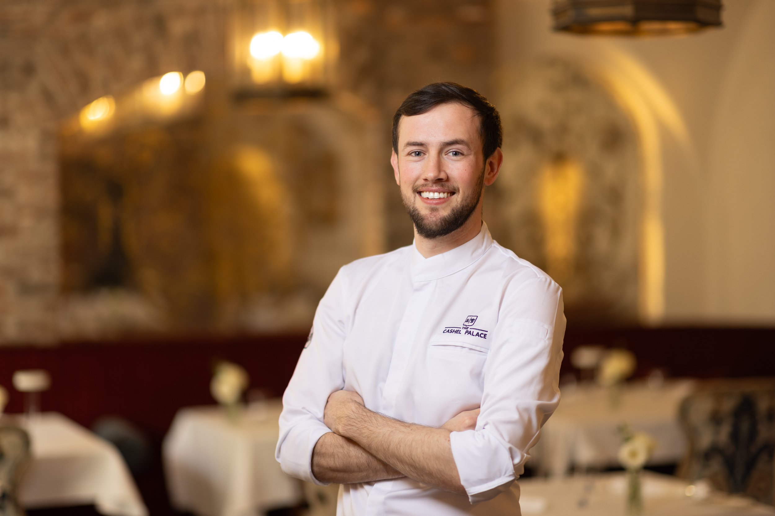 A male chef in a white uniform with the logo "The Cashel Palace" is smiling with arms crossed in a warmly lit restaurant or banquet hall with brick walls and blurred background.