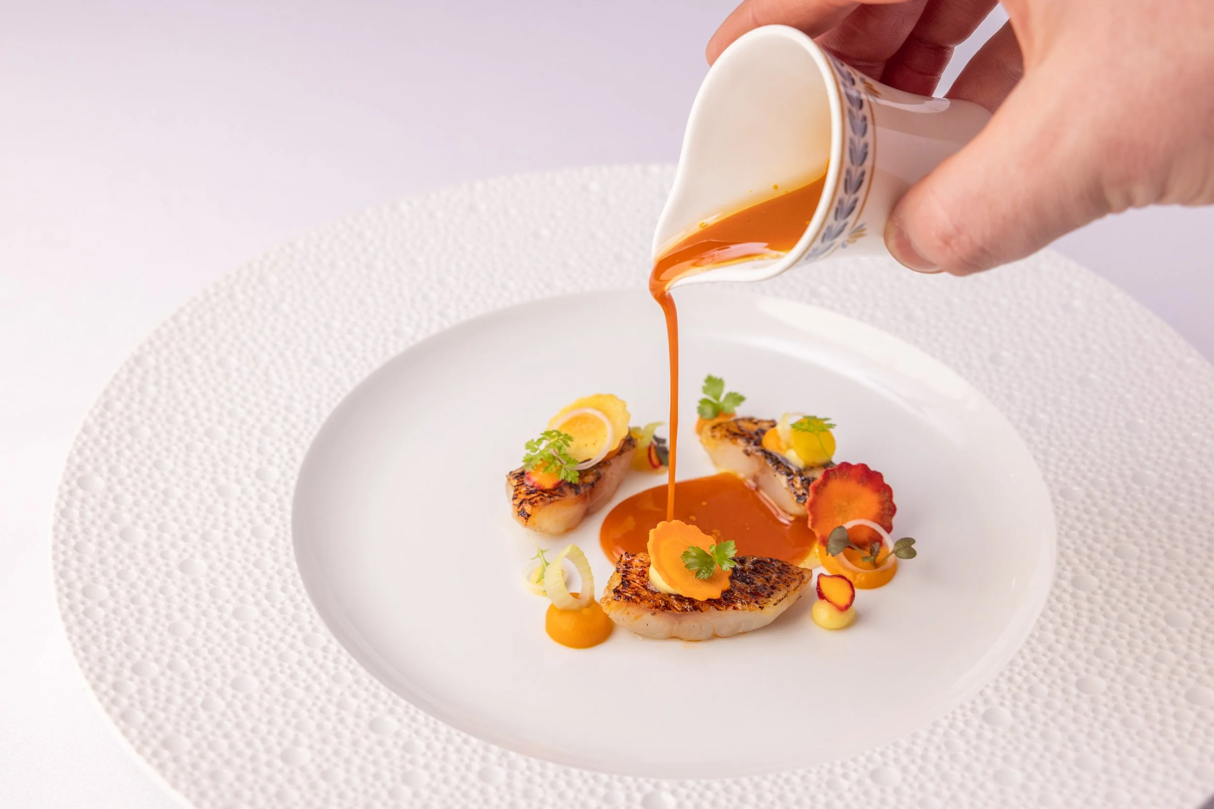 Chef pouring a brown sauce over a gourmet seafood dish with scallops and vegetables on a white textured plate.