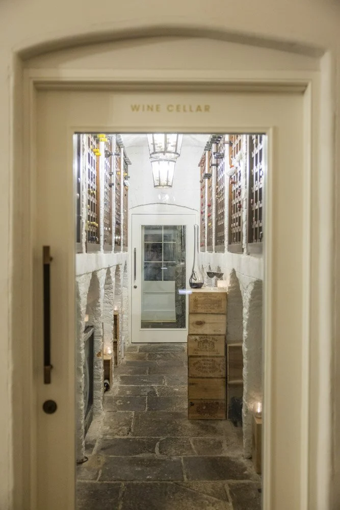 A view through the doorway of a wine cellar with stone walls, wooden crates, and wine bottles stored on racks. Candles are lit along the floor, creating a warm ambiance.