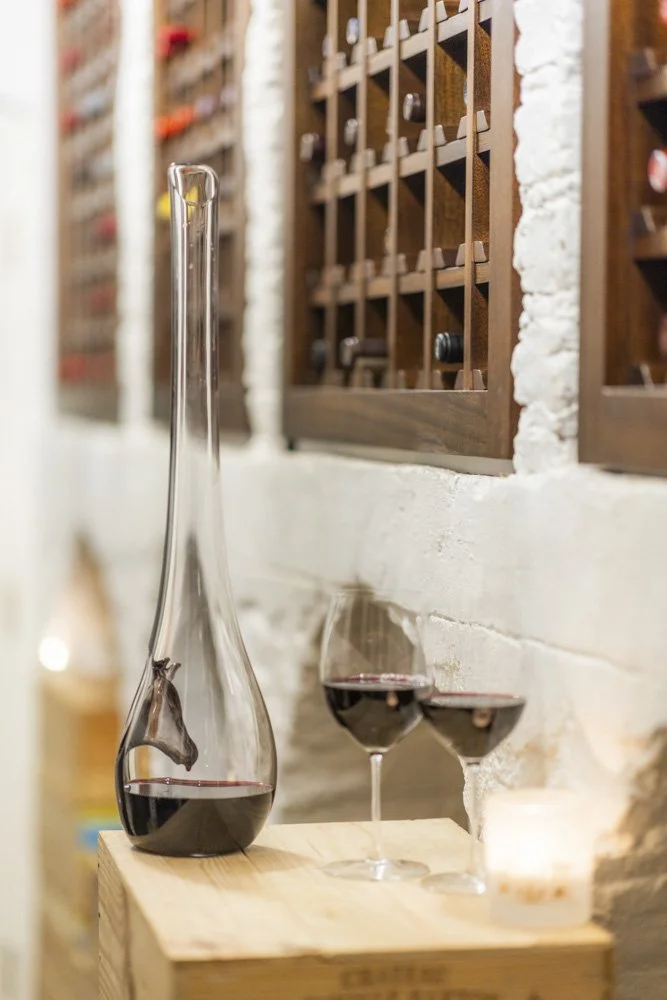 A wine decanter and two wine glasses filled with red wine on a wooden surface against a white brick wall with wine bottle cubby shelves.