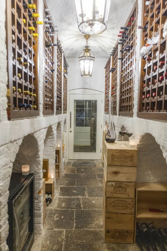 Wine cellar with stone walls and floor, wooden wine racks on both sides, candles, and a glass door at the end.
