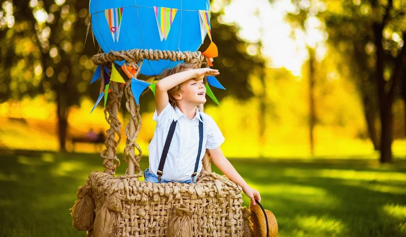 Young boy in a white shirt and suspenders standing in a hot air balloon basket decorated with colorful bunting flags, holding a hat in one hand and shading his eyes with the other, looking into the distance in a park on a sunny day.