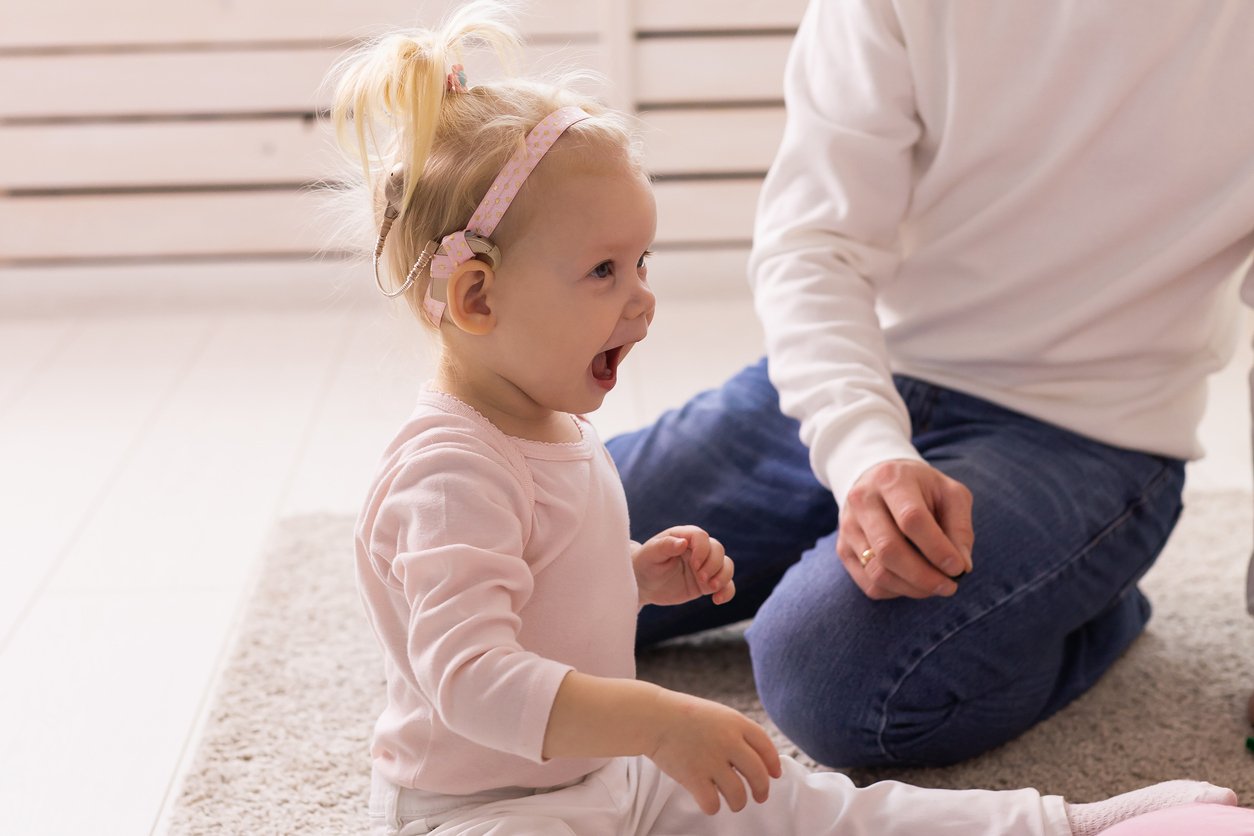 A young girl with a cochlear implant, sitting on a light-colored carpet with an excited expression after learning to hear new sounds during speech therapy for aural rehabilitation in Knoxville, TN.