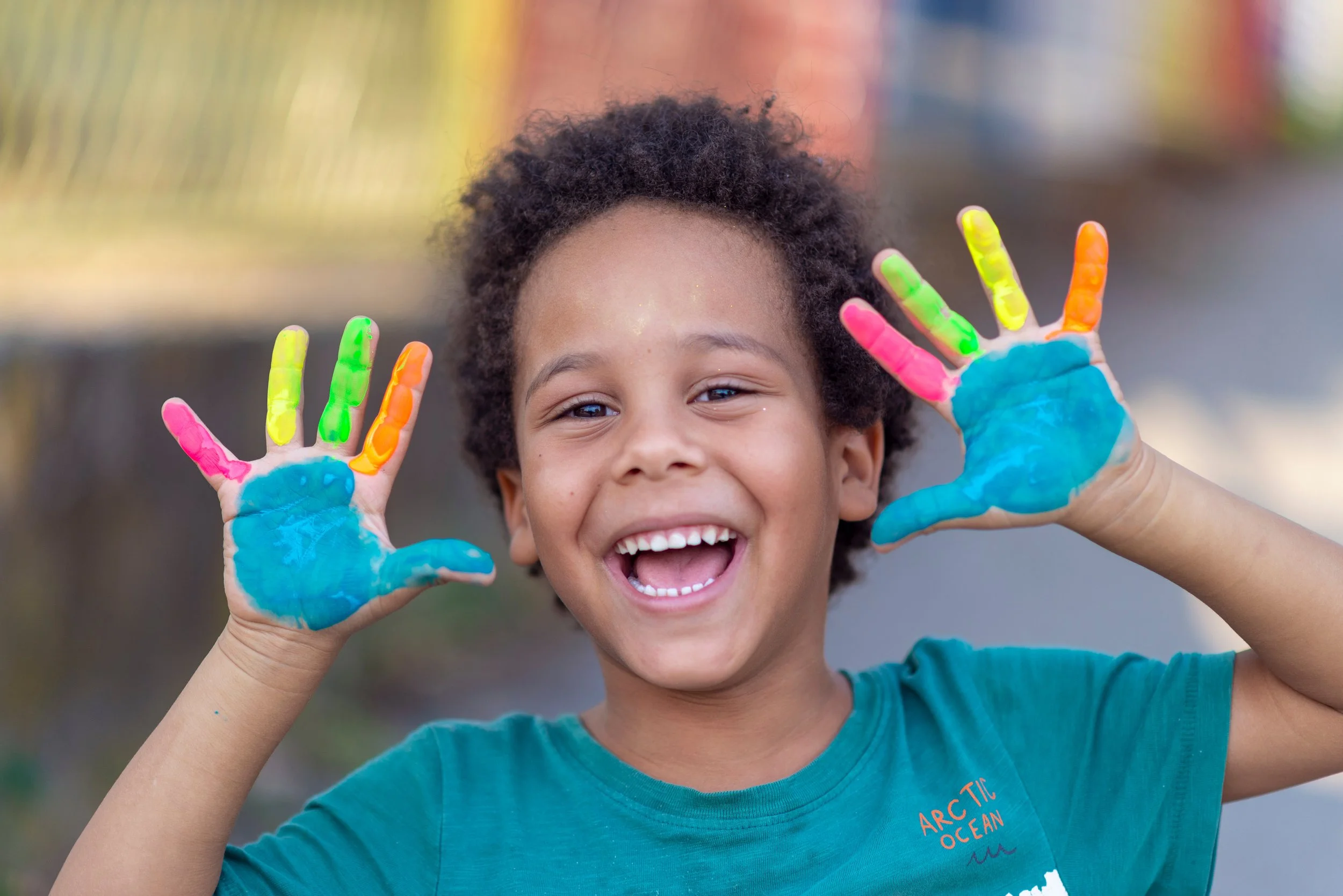 A smiling young child with curly hair is holding up both hands covered in colorful paint during play-based speech therapy for kids in Knoxville, TN