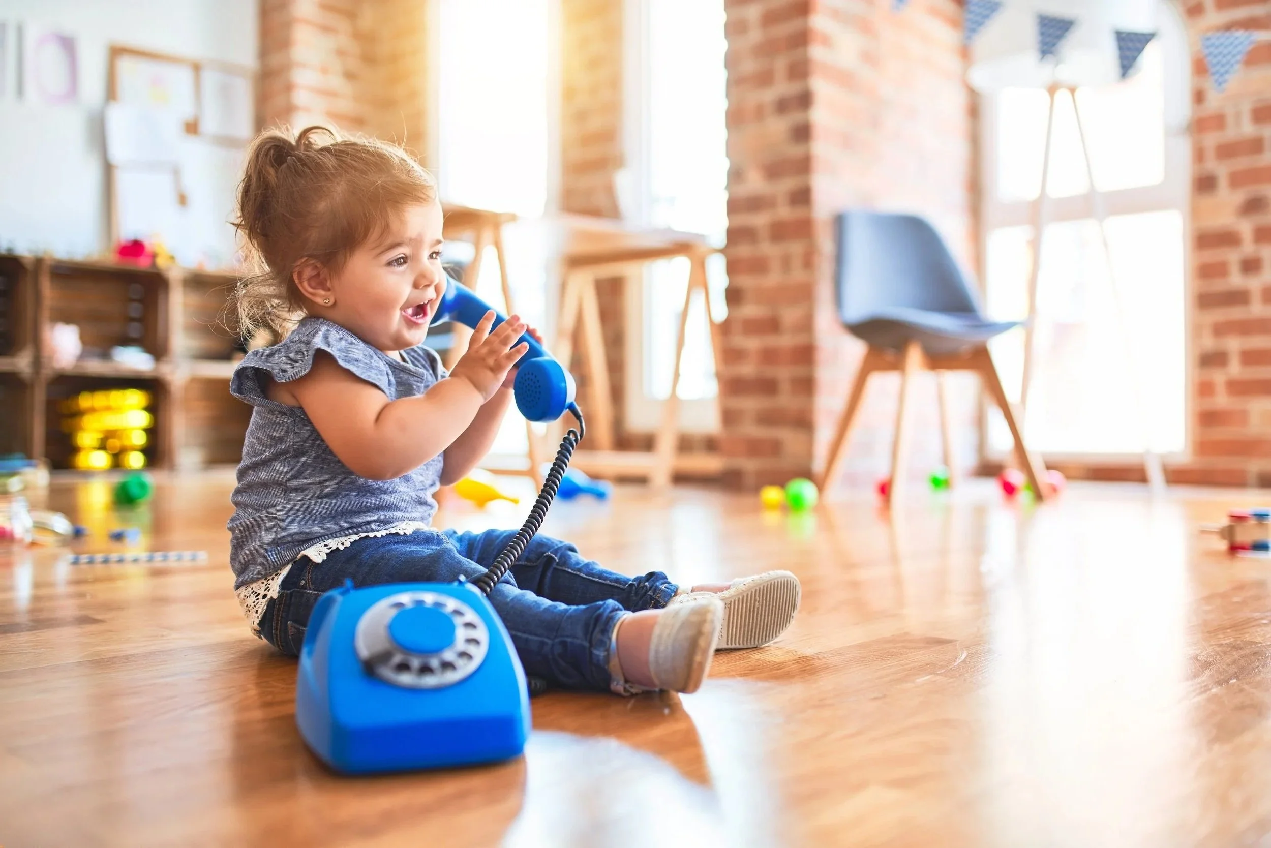 A young girl sitting on the wooden floor playing with a blue toy telephone, working on listening and talking during speech therapy for children in Knoxville, TN
