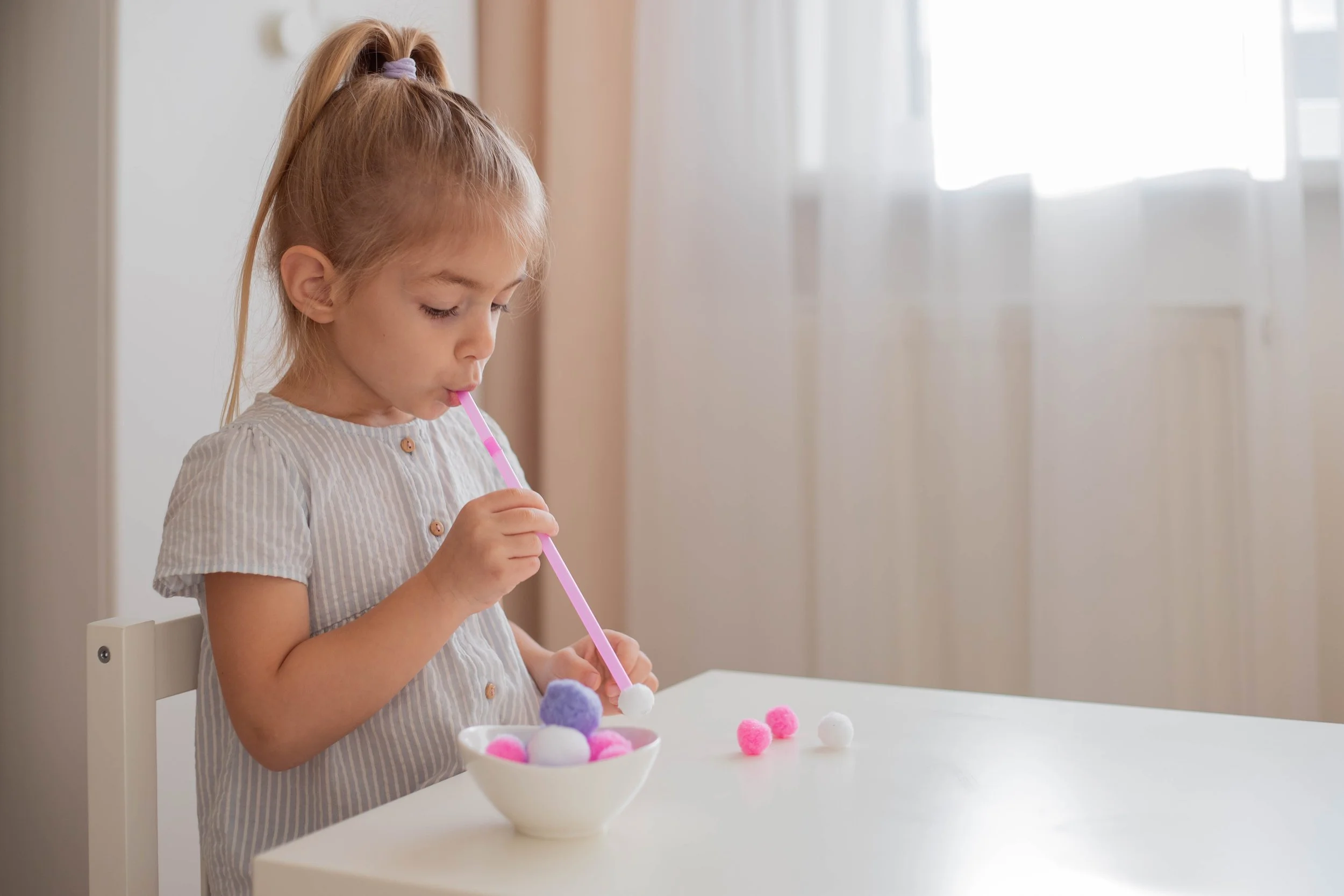 A young girl with a blonde ponytail sucking through a straw to move pom-poms to a bowl, working on her oral motor skills during myofunctional therapy for kids in Knoxville, TN.