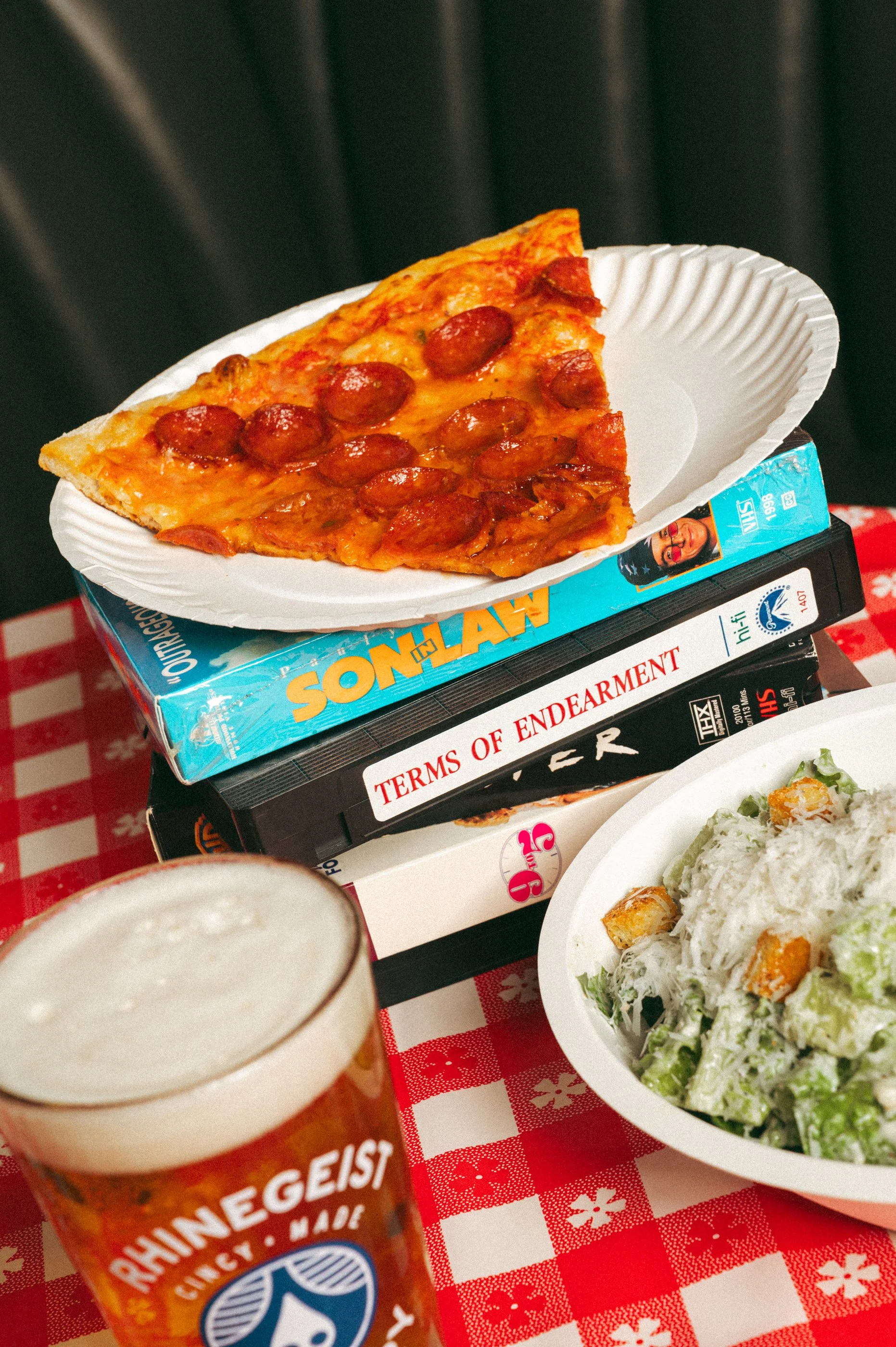 A slice of pepperoni pizza on a white paper plate, placed on top of a pile of books. A glass of beer and a bowl of salad are also visible on a red and white checkered tablecloth.