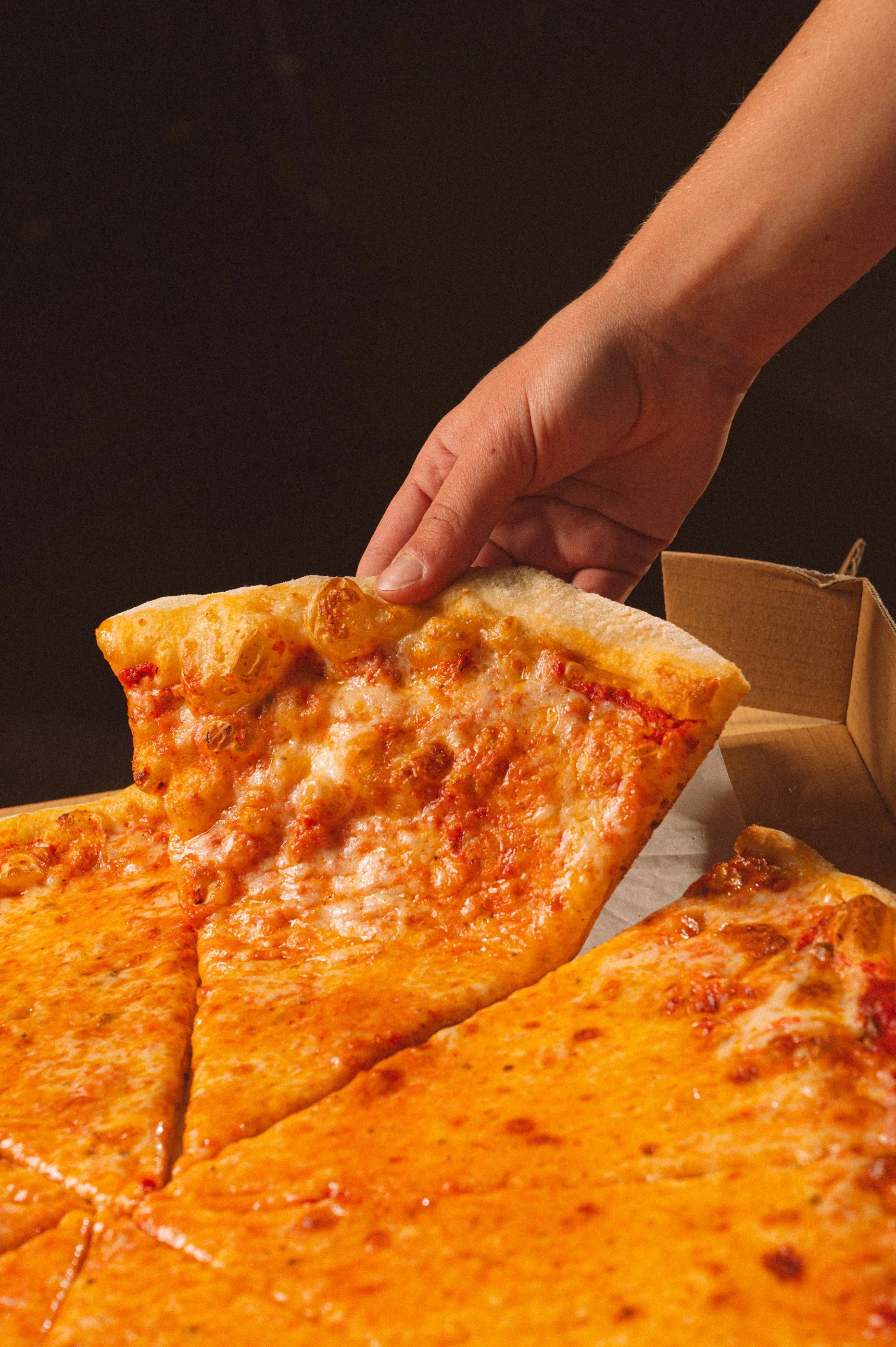 Close-up of a hand picking up a slice of cheese and pepperoni pizza from a box, showing several other slices of cheese pizza.
