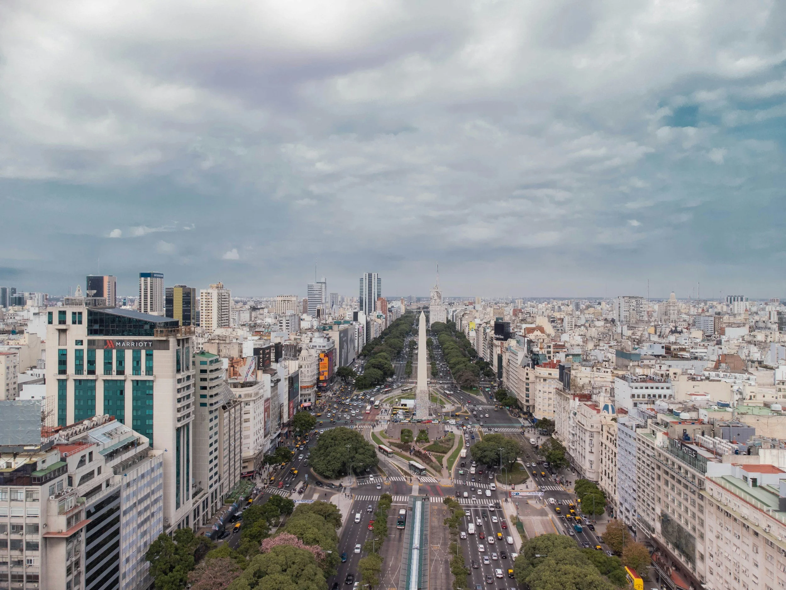 A city skyline featuring tall buildings, wide streets with cars and buses, a park with trees and a fountain, and a cloudy sky overhead.