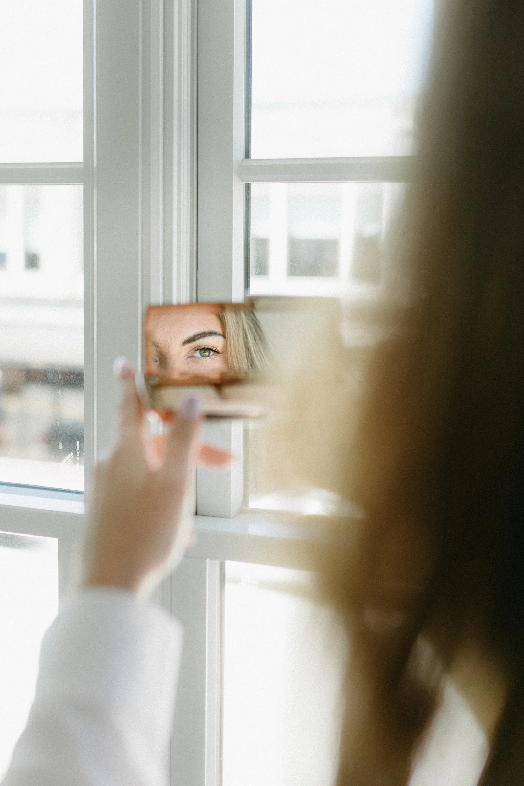 Person taking a selfie in front of a mirror, focusing on their eye, with windows in the background.