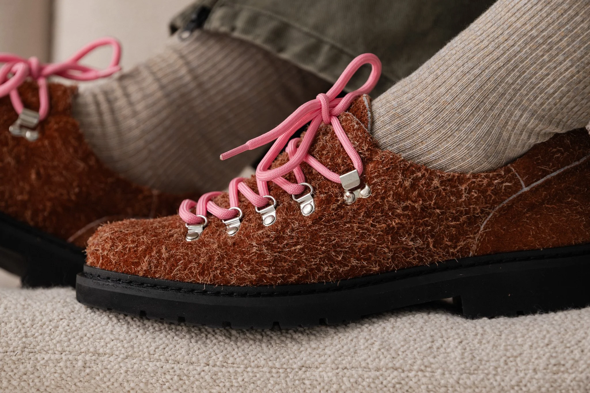 Close-up of a brown hiking boot with pink laces, seen on a person's foot, with tan and green pants, resting on a beige carpet.