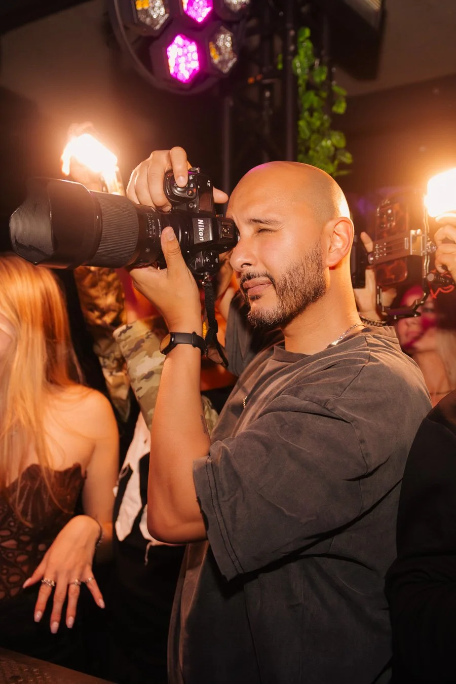 A man with a bald head and beard is taking a photo using a professional camera at what appears to be a lively event or party with colorful lighting and several people around, some wearing masks.