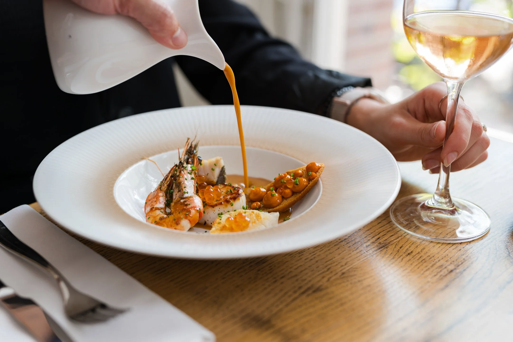 Person pouring brown sauce over a fancy seafood dish with shrimp, white fish, and a slice of toasted bread topped with orange caviar and herbs on a white plate.
