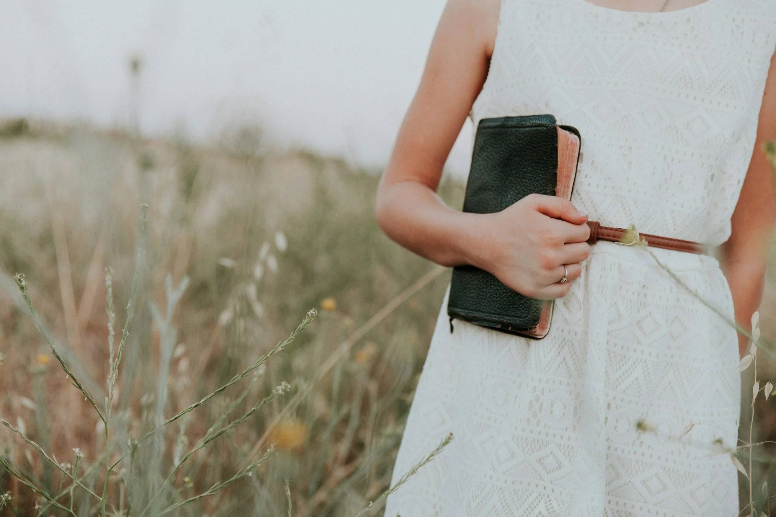 Person wearing a white dress holding a black and brown notebook or wallet in a field of tall grasses.