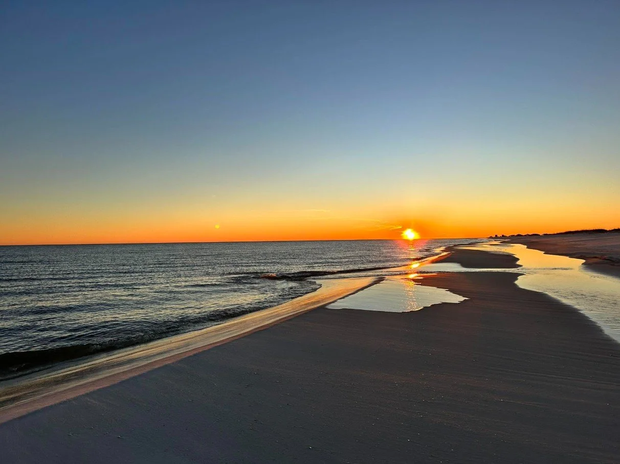 Sunset over the ocean with a sandy beach and small water pools reflecting the sky, calm waves, and a clear sky transitioning from blue to orange.