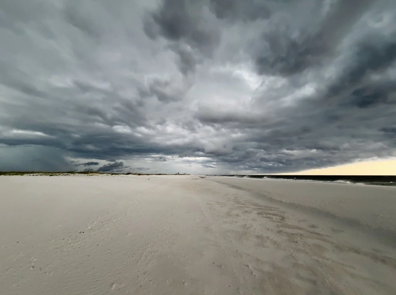 A dramatic storm sweeping across an empty white-sand beach, with dark clouds over the shoreline and a narrow break of light on the horizon.